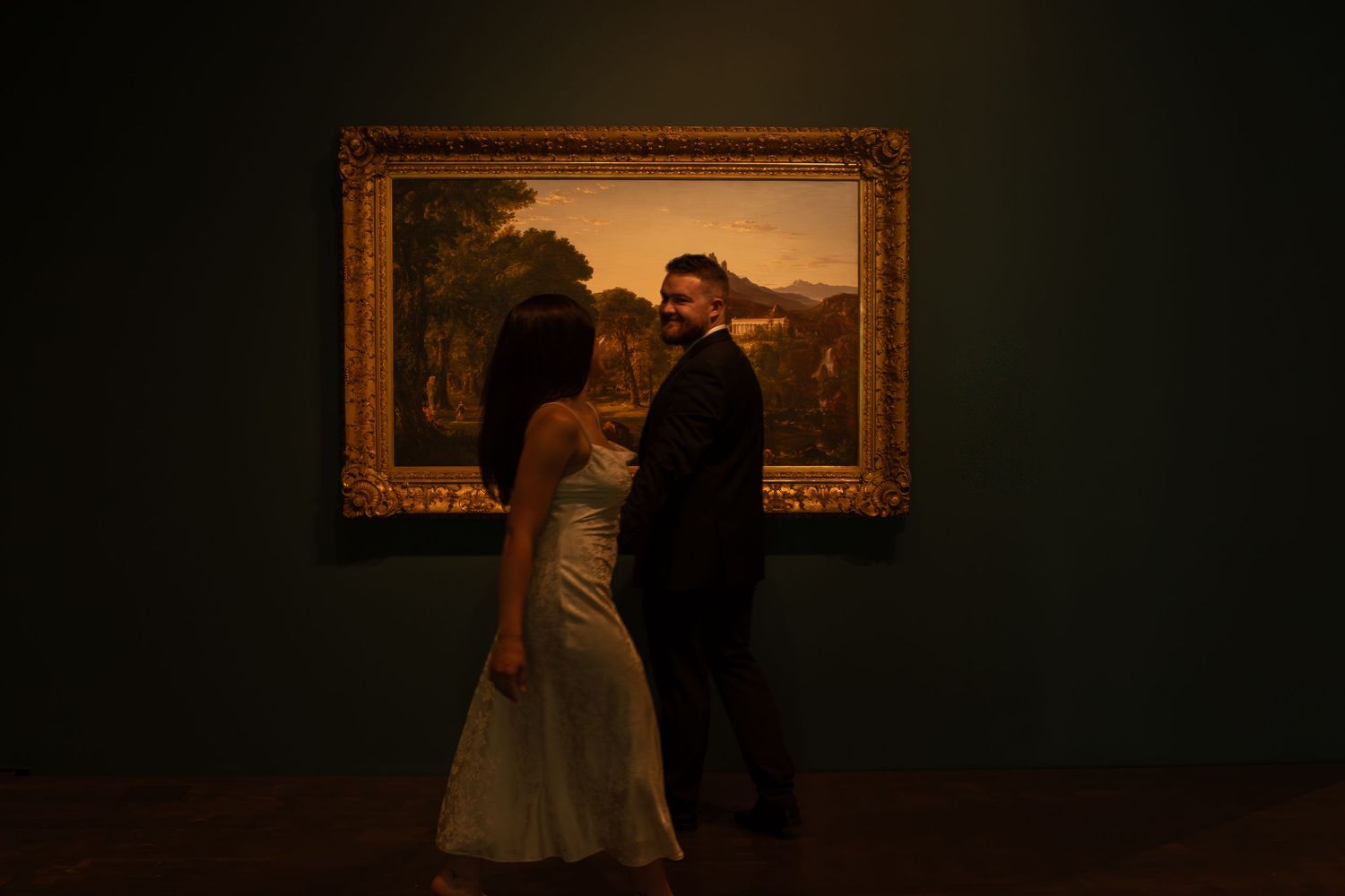 Two silhouettes examine framed artwork on a darkly lit gallery wall.