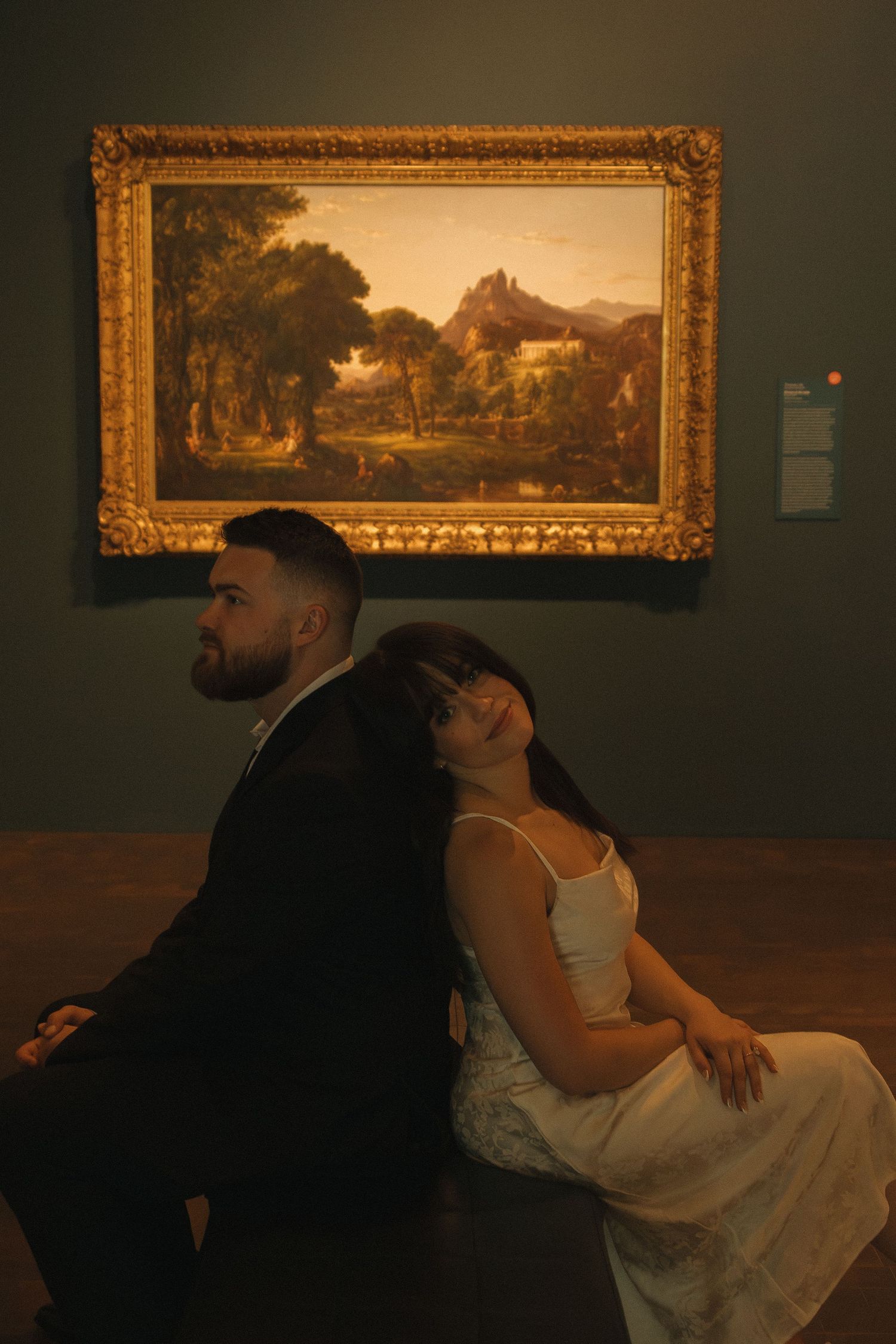 Couple sits back-to-back in front of framed landscape painting in museum.