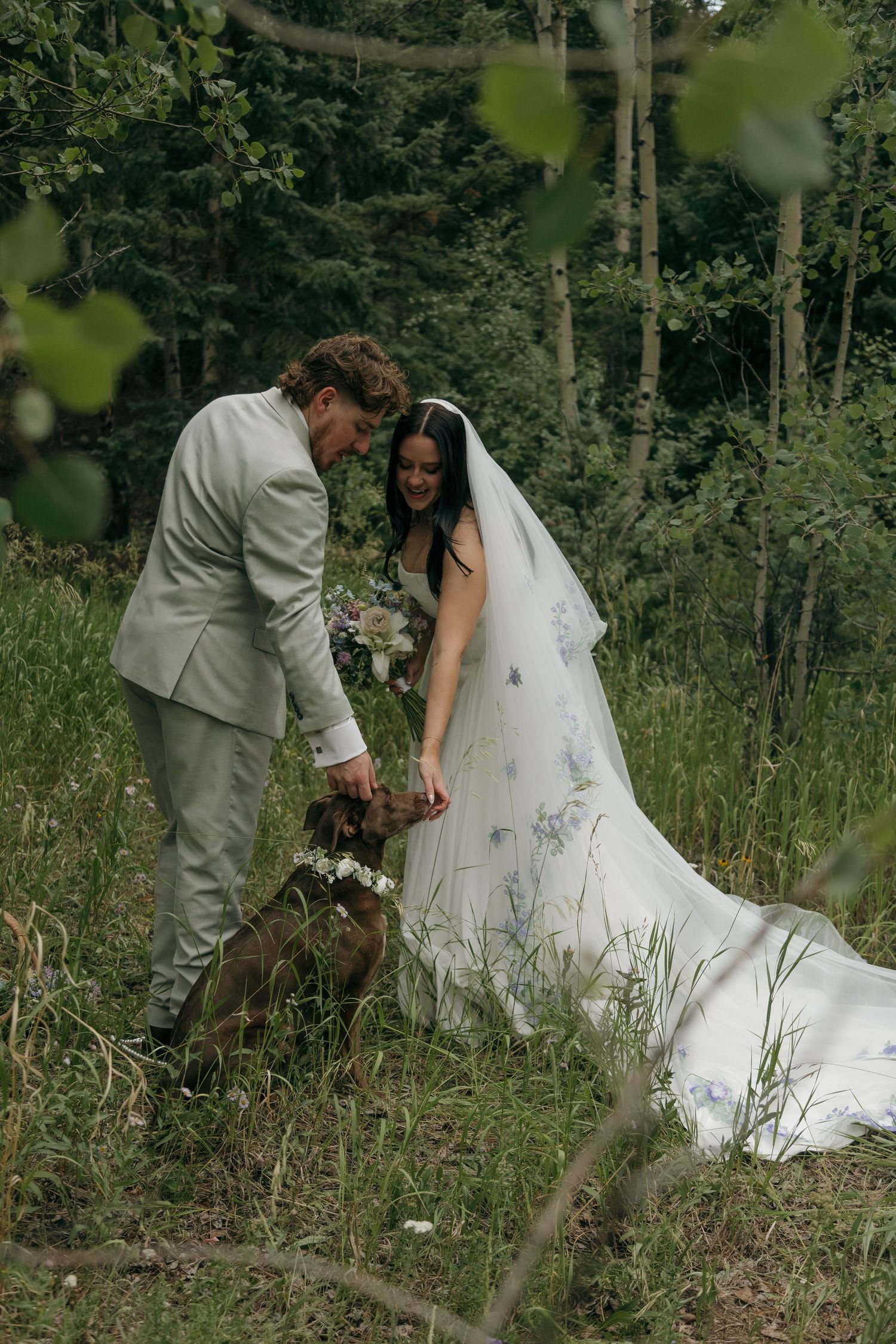 Bride and groom in forest setting petting their dog during wedding photos, bride wearing floral veil.