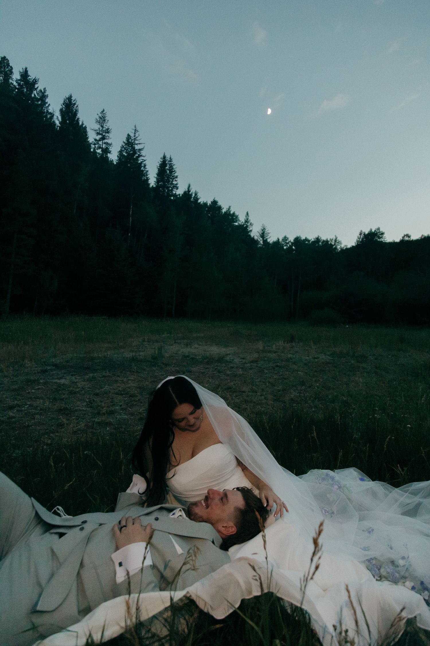 Bride leaning over groom lying in grass at dusk, crescent moon visible in dusky sky.