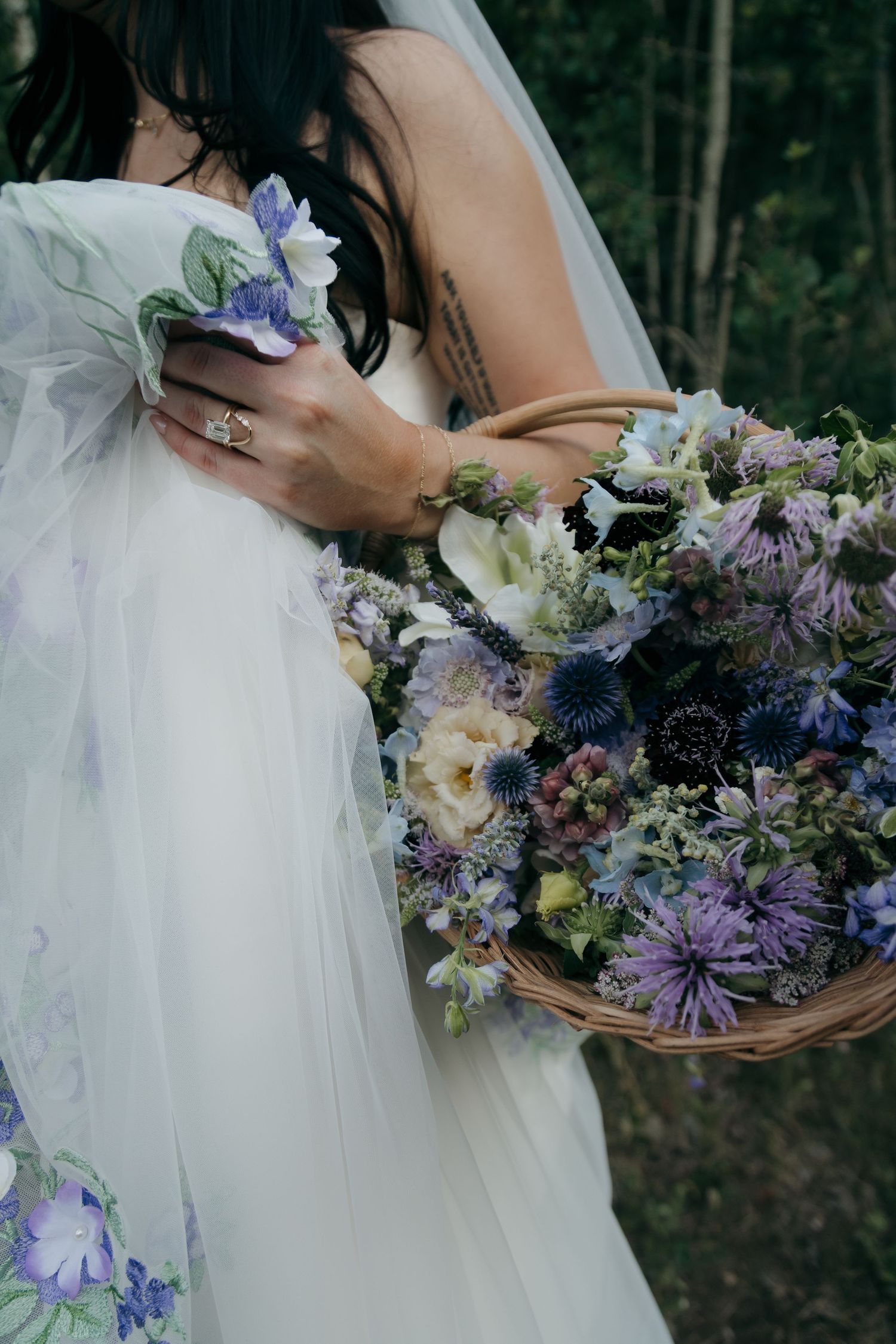 Bride holds a wicker basket bouquet of purple and blue wildflowers, wearing a floral-embroidered gown.