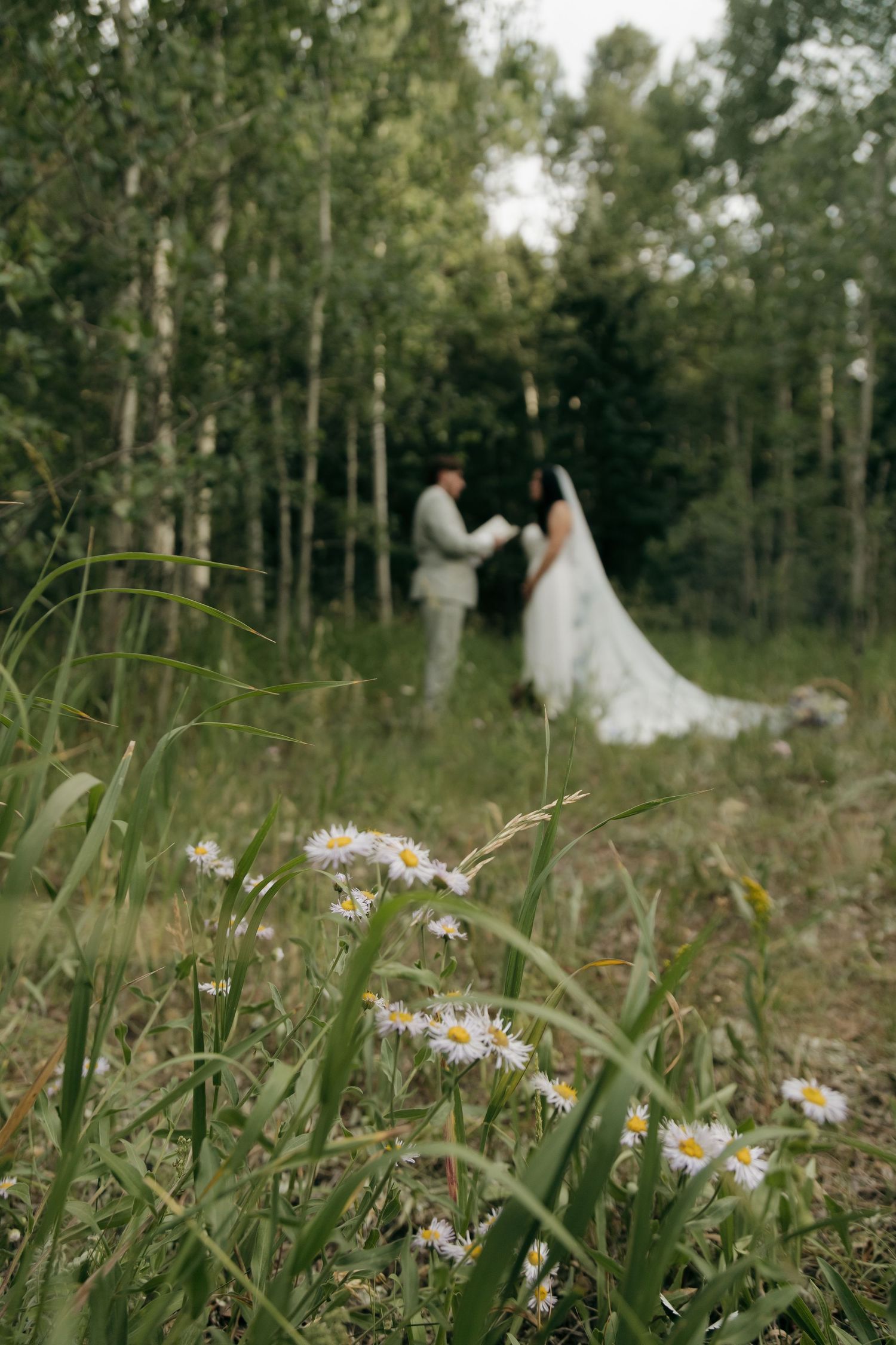 Wildflowers frame blurred bride and groom holding hands in a green forest meadow during outdoor wedding ceremony.