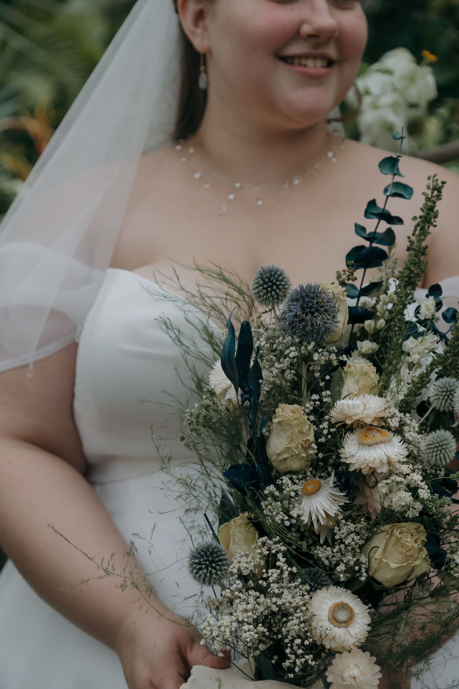 Bride in white off-shoulder gown holding a rustic bouquet with blue thistles, white roses, and eucalyptus.