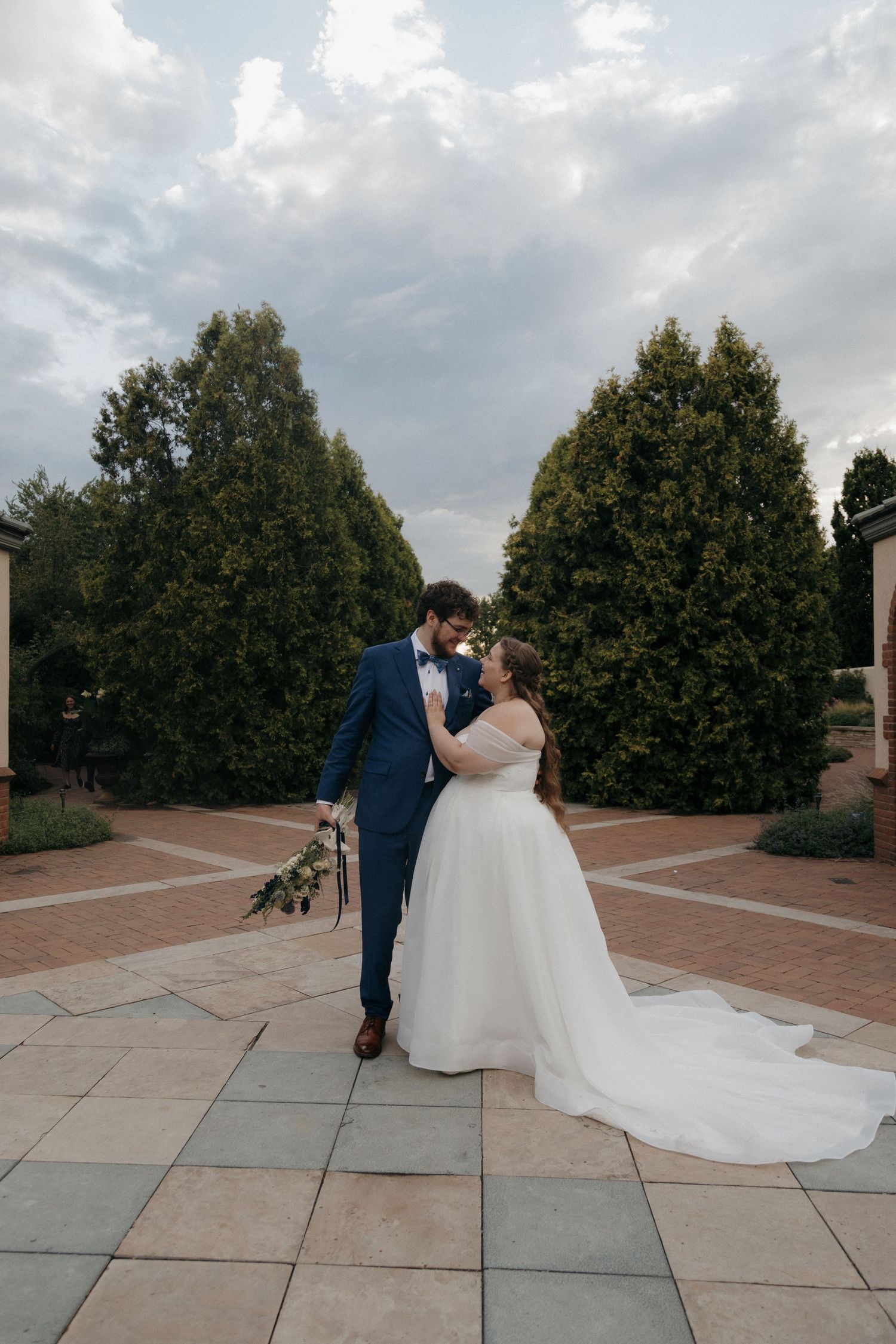 Bride and groom share romantic kiss on brick courtyard surrounded by tall green trees at dusk.