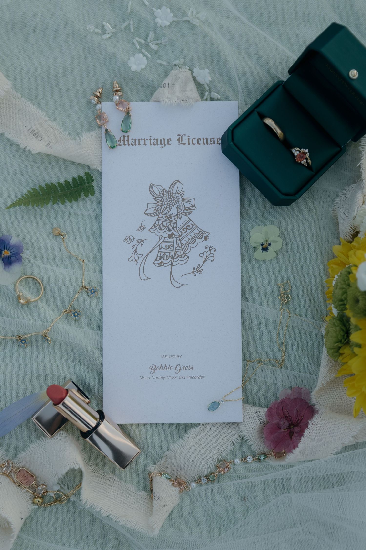 Elegant wedding flat lay with marriage license, rings in teal box, jewelry, lipstick, and wildflowers on sage green fabric.