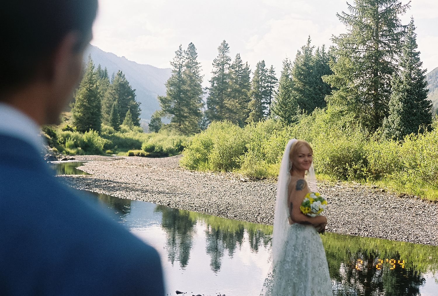 Bride in white gown holding bouquet stands by mountain stream as groom watches.
