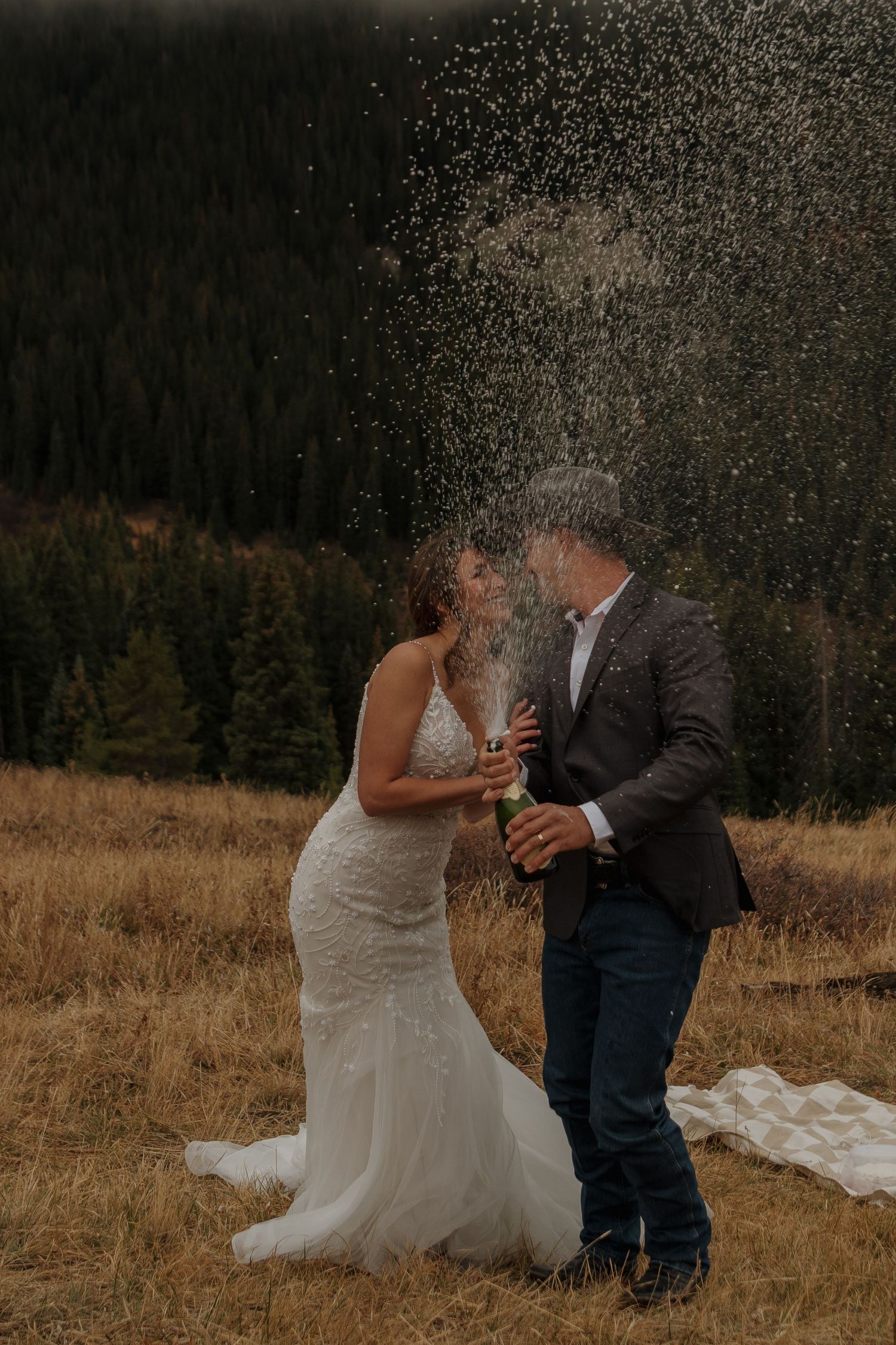 A couple celebrates with champagne spray in a mountain meadow setting.