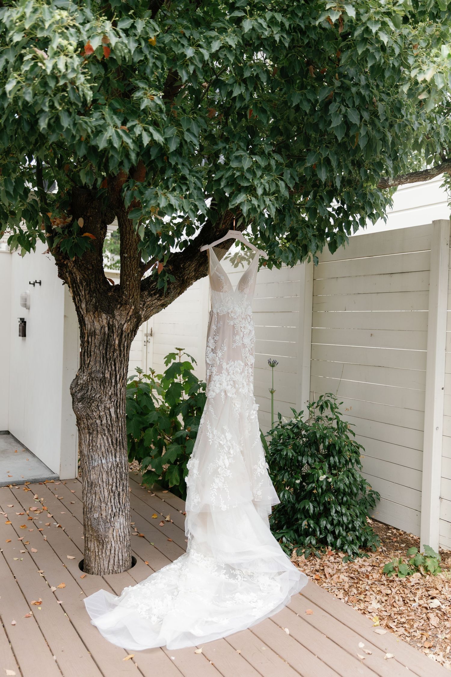 A white wedding dress hangs elegantly from a tree branch against a light-colored wall backdrop.