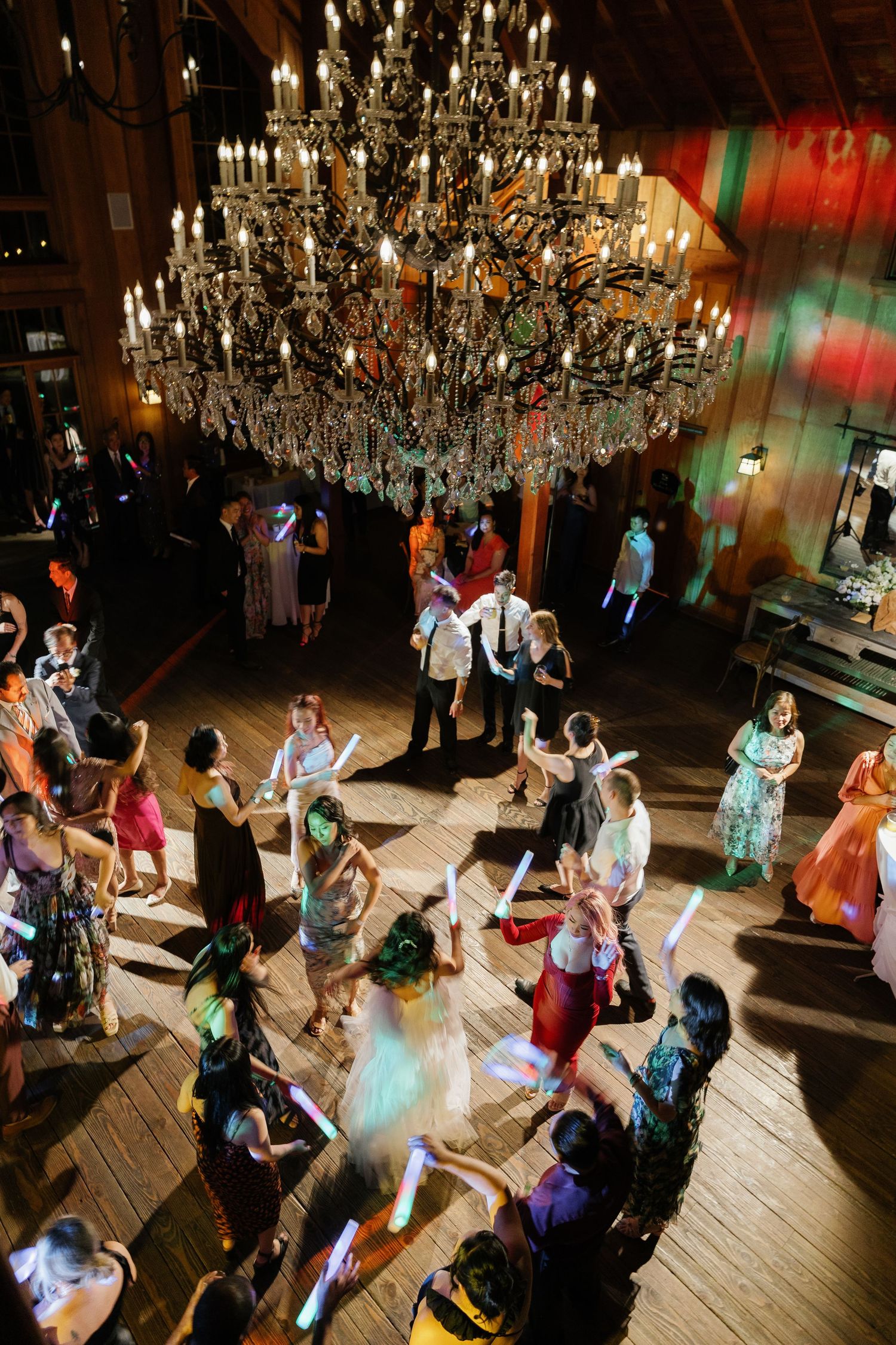 Wedding guests dance under an ornate crystal chandelier at a reception venue with colorful lighting and wooden floors.