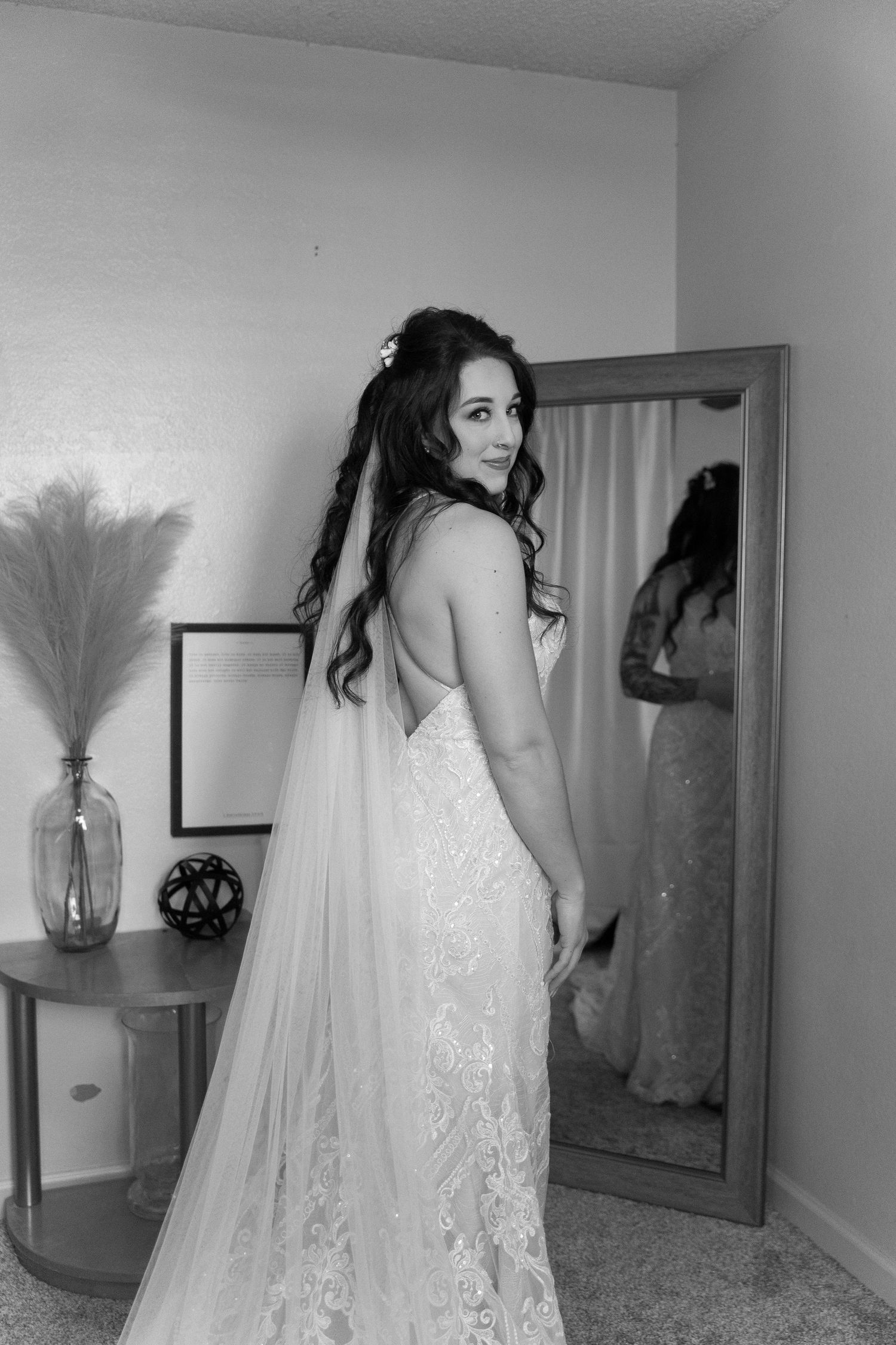 Person in lace wedding dress and long veil poses beside full-length mirror.