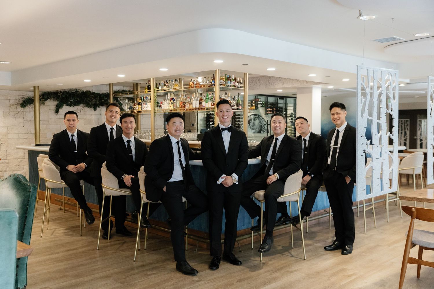 Groomsmen in black suits pose creatively in a modern indoor setting with white chairs and wood flooring.