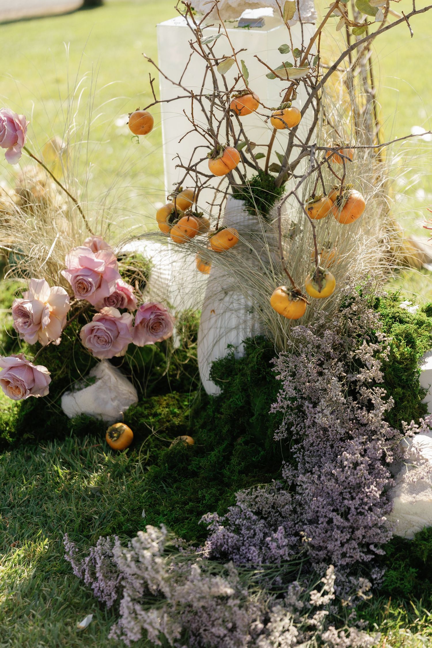 Elegant floral arrangement featuring pink roses, orange persimmons, and delicate purple flowers against a grassy backdrop.