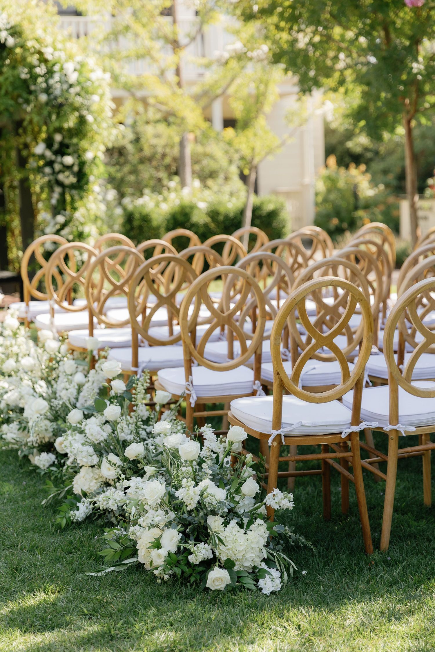 Elegant wooden crossback chairs with white cushions arranged in rows on a lush green lawn for an outdoor wedding ceremony.