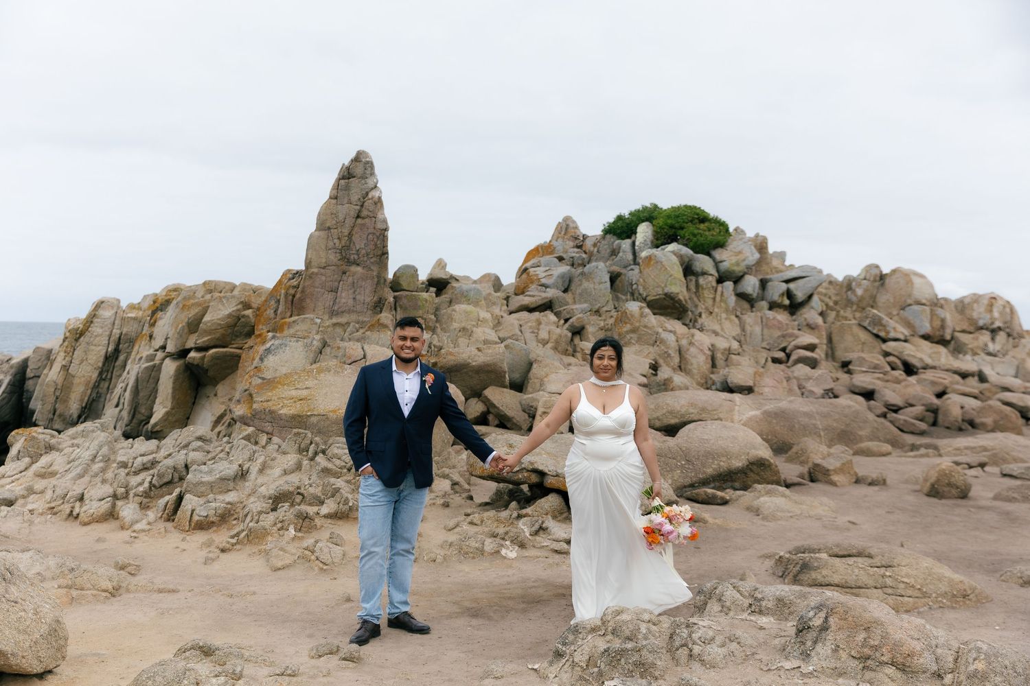 Wedding couple posing on rocky coastal cliffs with ocean and beach views in the background.