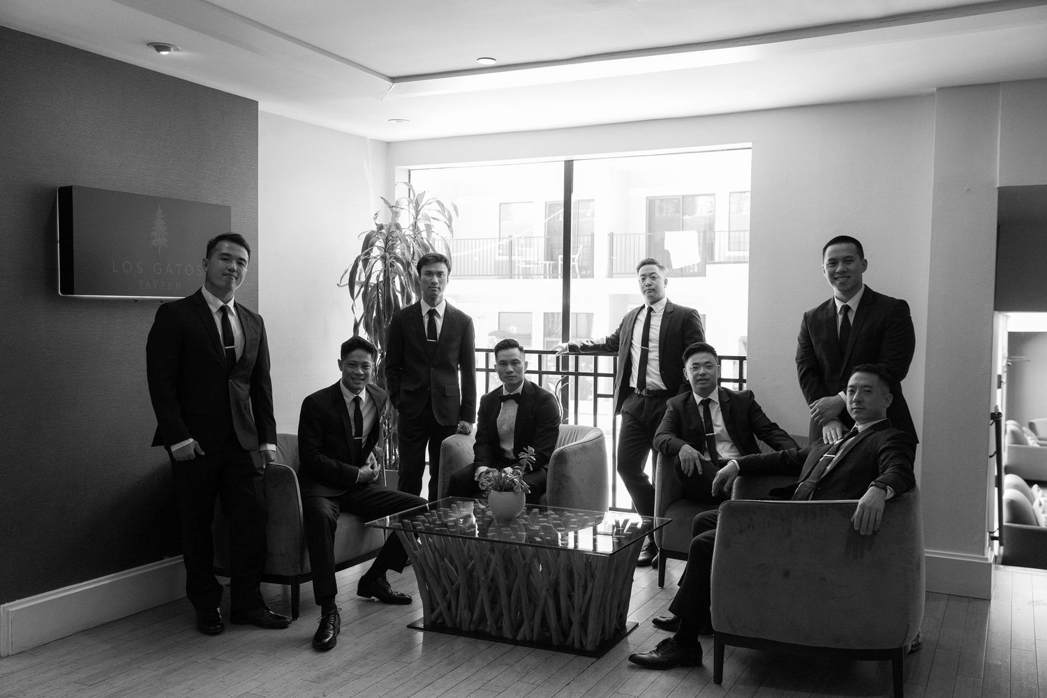 Wedding party members gather in a bright modern room with large windows and comfortable seating.