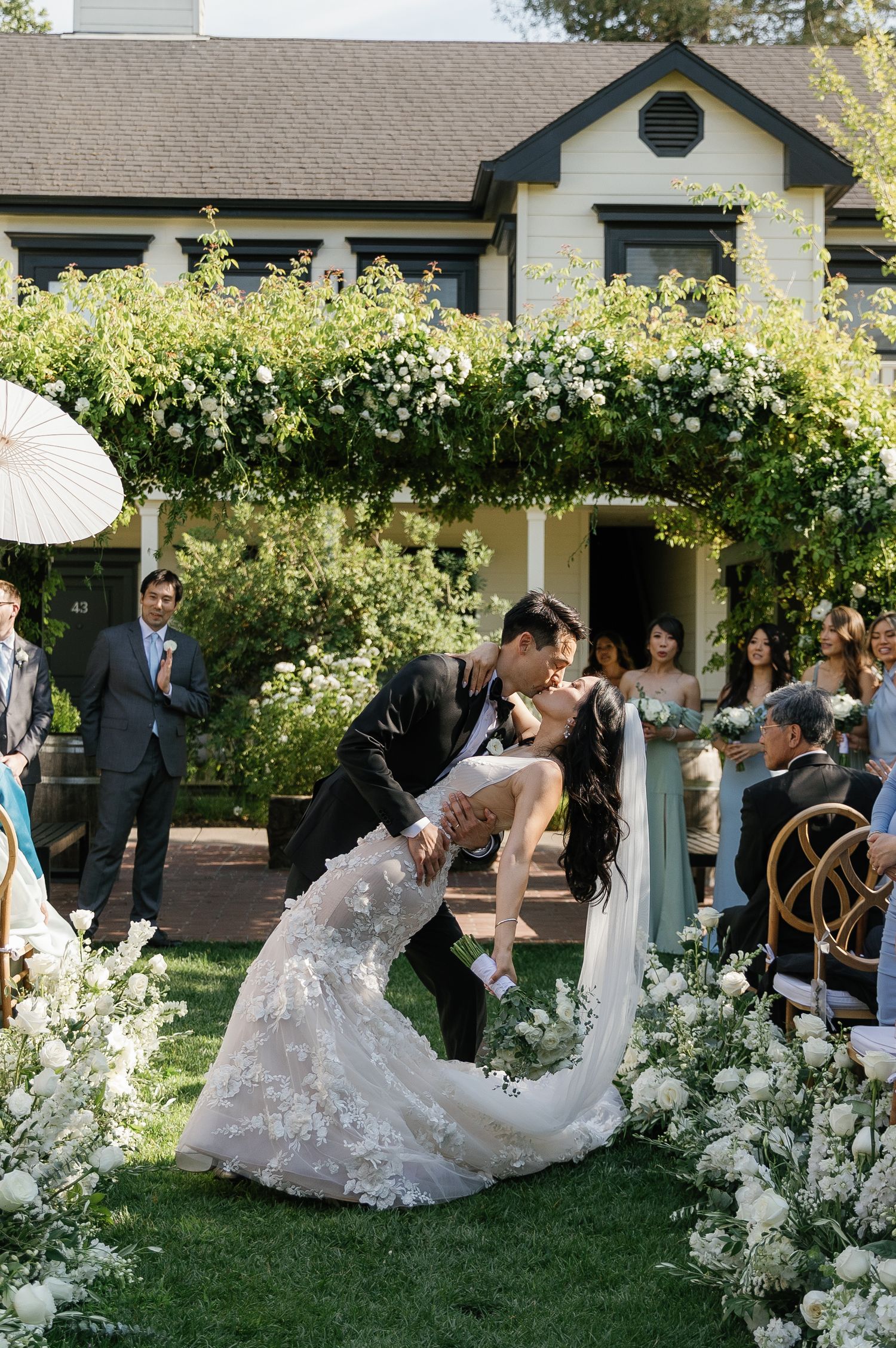 A wedding party dances and celebrates on a garden lawn surrounded by white floral arrangements and greenery.
