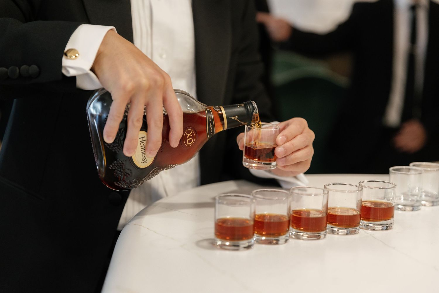 A bartender pours multiple shots of amber-colored liquor into small glasses lined up on a bar counter.