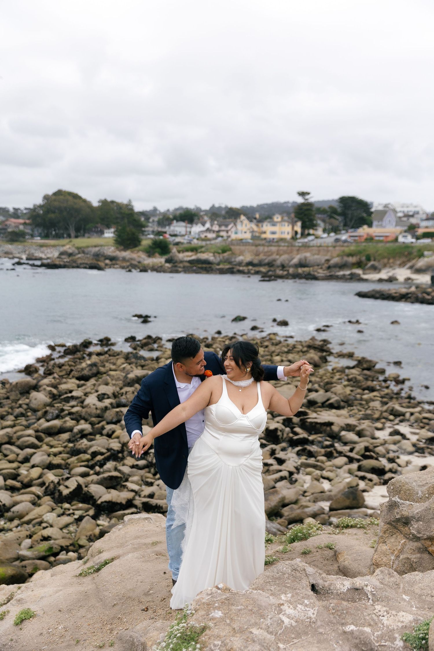 A couple shares a romantic moment on the rocky New England coastline with boats and houses visible in the background.