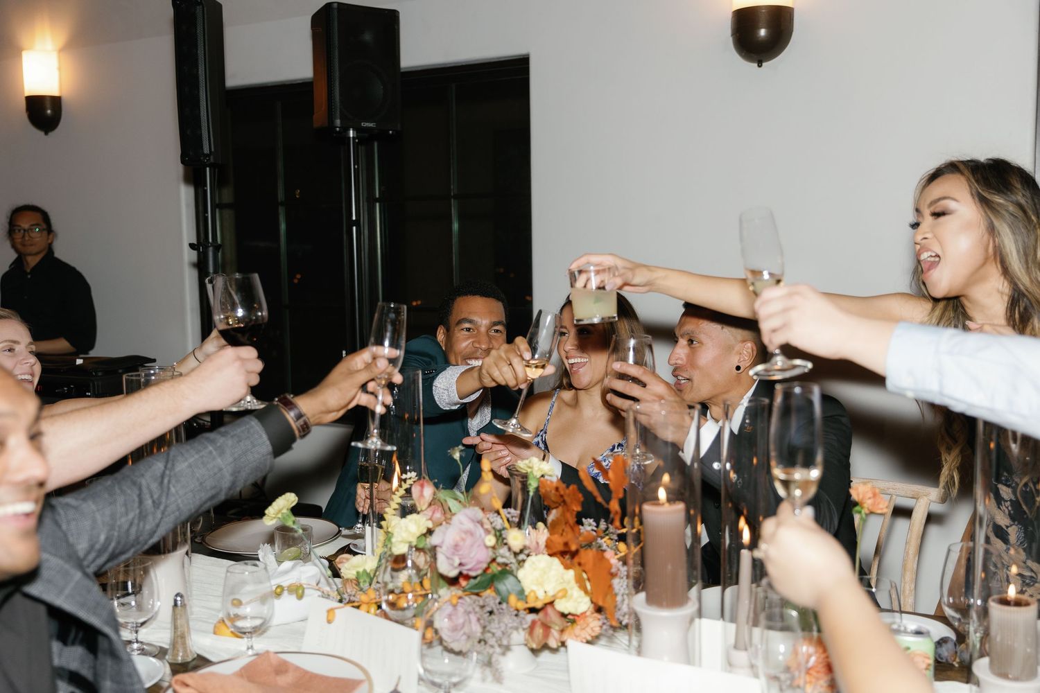 People raise their arms in celebration at a lively dinner party with tables set for a festive gathering.