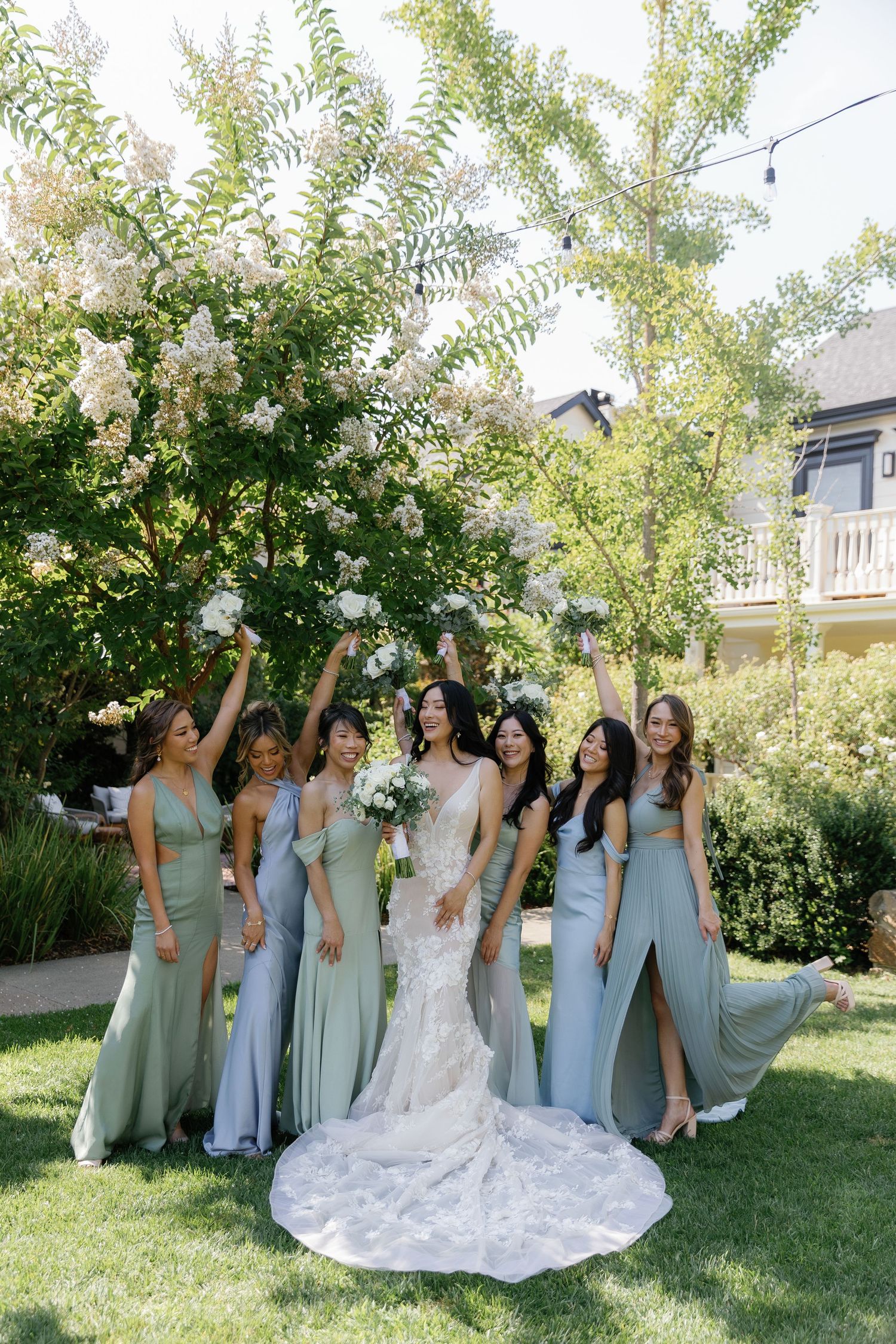 A wedding party in sage green bridesmaid dresses poses together on a sunny lawn for outdoor photos.
