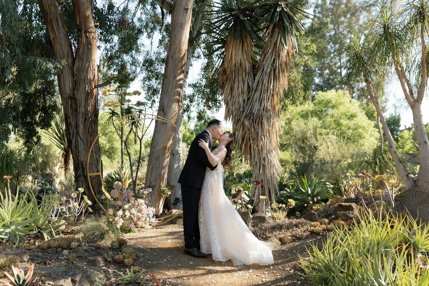 A wedding couple shares an intimate moment among tall palm trees and lush garden landscaping.