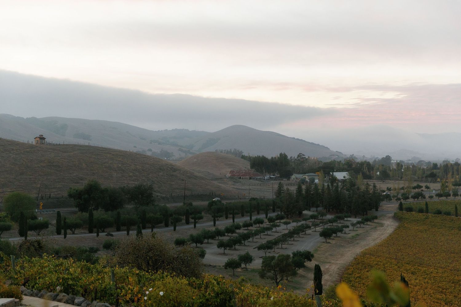 Scenic panoramic view of rolling hills and mountains at sunset with scattered buildings and winding roads in the distance.