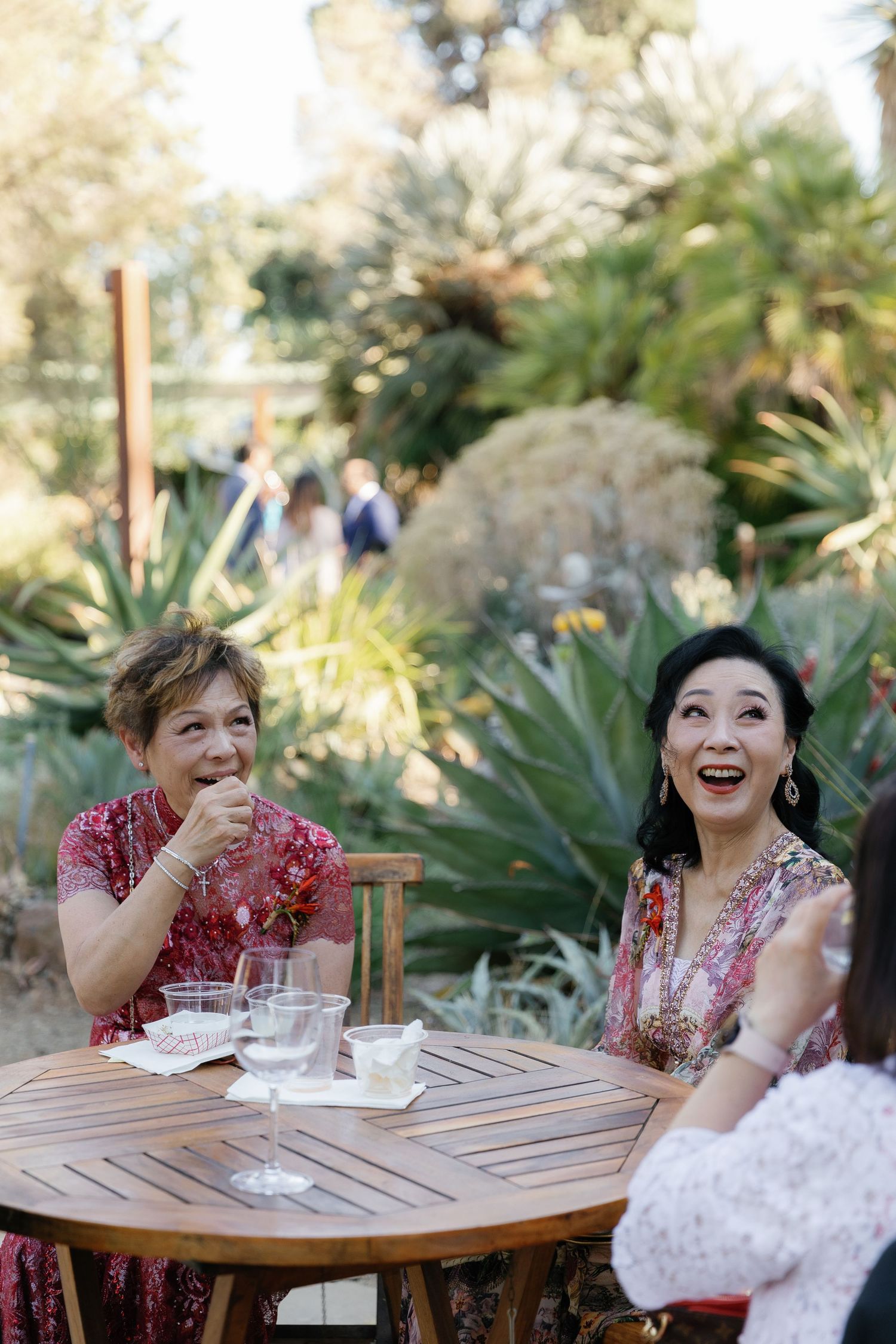 People sharing laughter and conversation while seated at a wooden table in a sunny garden setting.