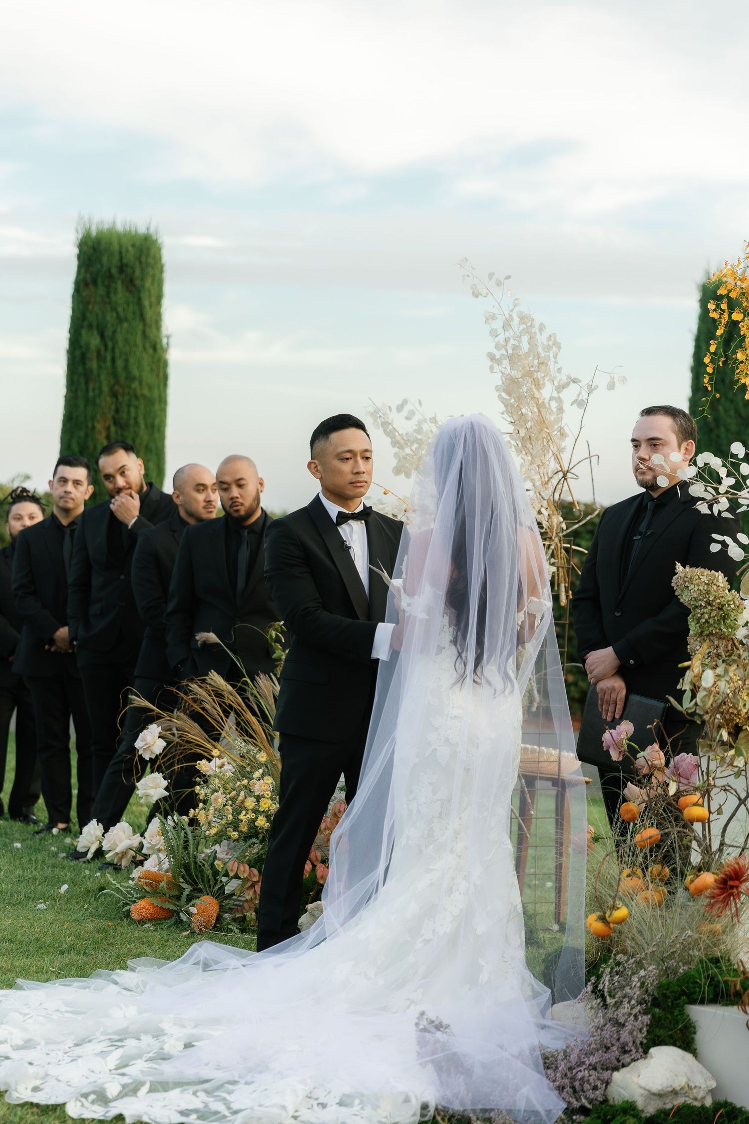 Wedding ceremony guests gather outdoors around a bride in a flowing white veil and dress amid autumn decorations.