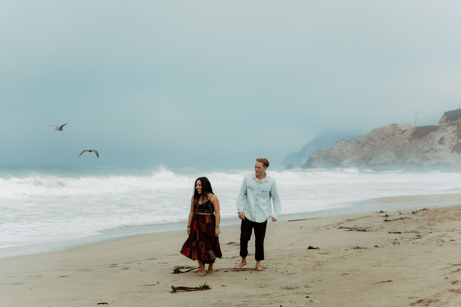 A couple walks together along a misty beach with crashing waves and seagulls flying overhead near rocky cliffs.