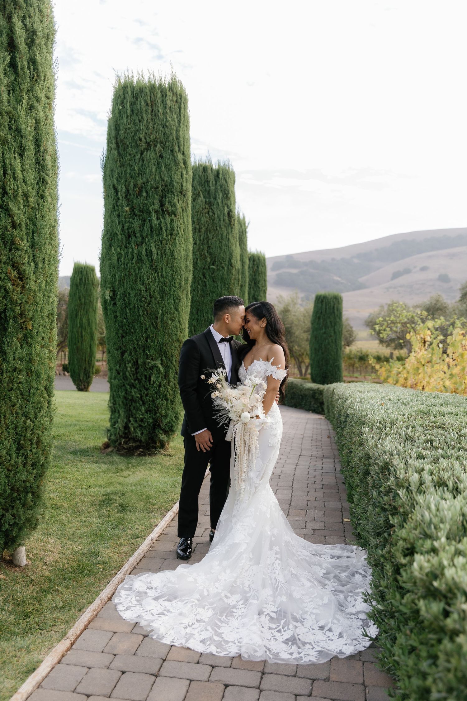 Romantic wedding photoshoot in a garden path lined with tall cypress trees and mountain views.