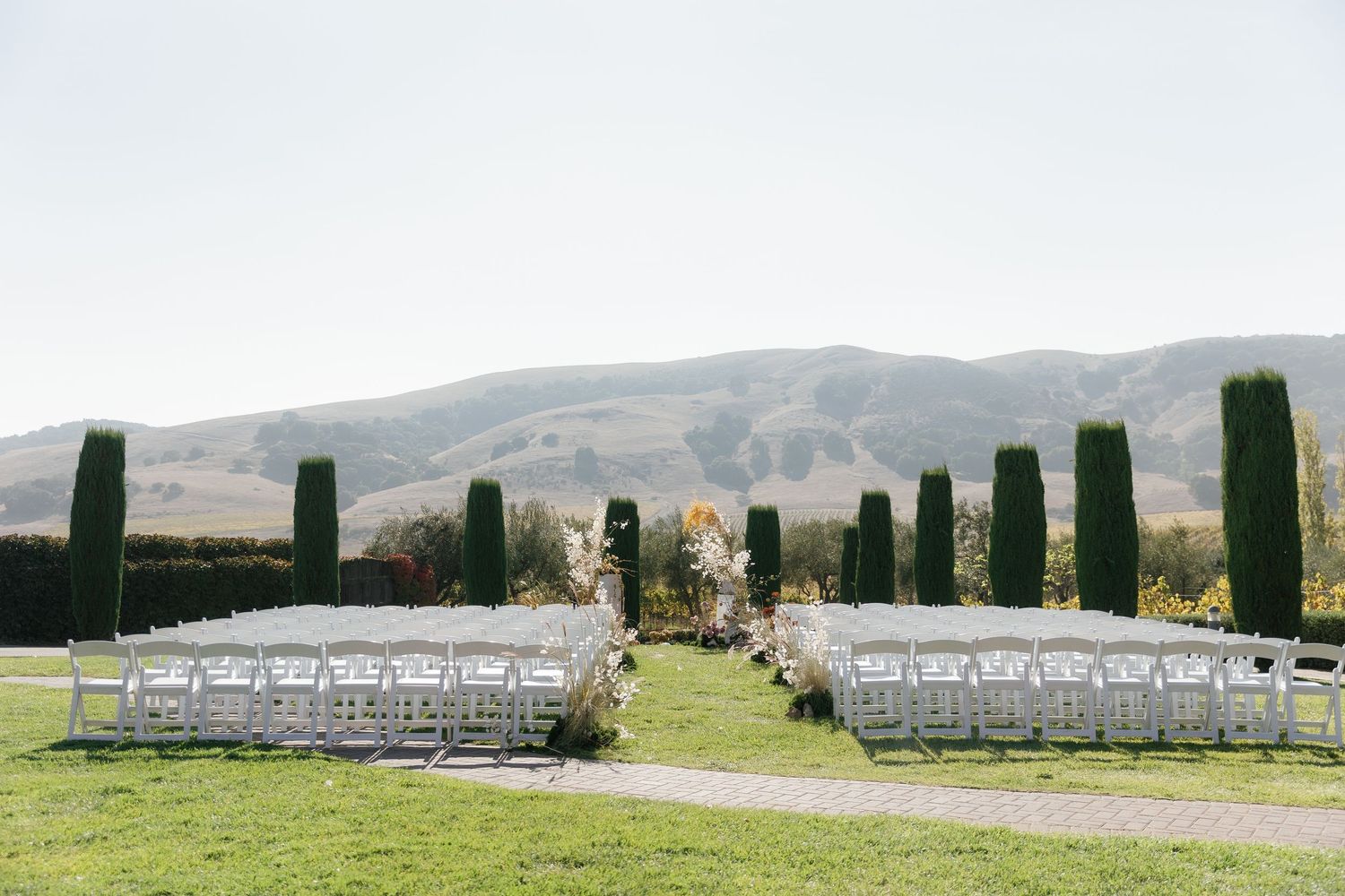 White chairs arranged in rows for an outdoor wedding ceremony with rolling hills and cypress trees in the background.