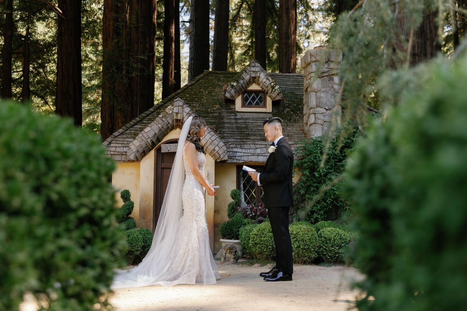A wedding couple stands together on a stone path surrounded by lush greenery and garden landscaping.