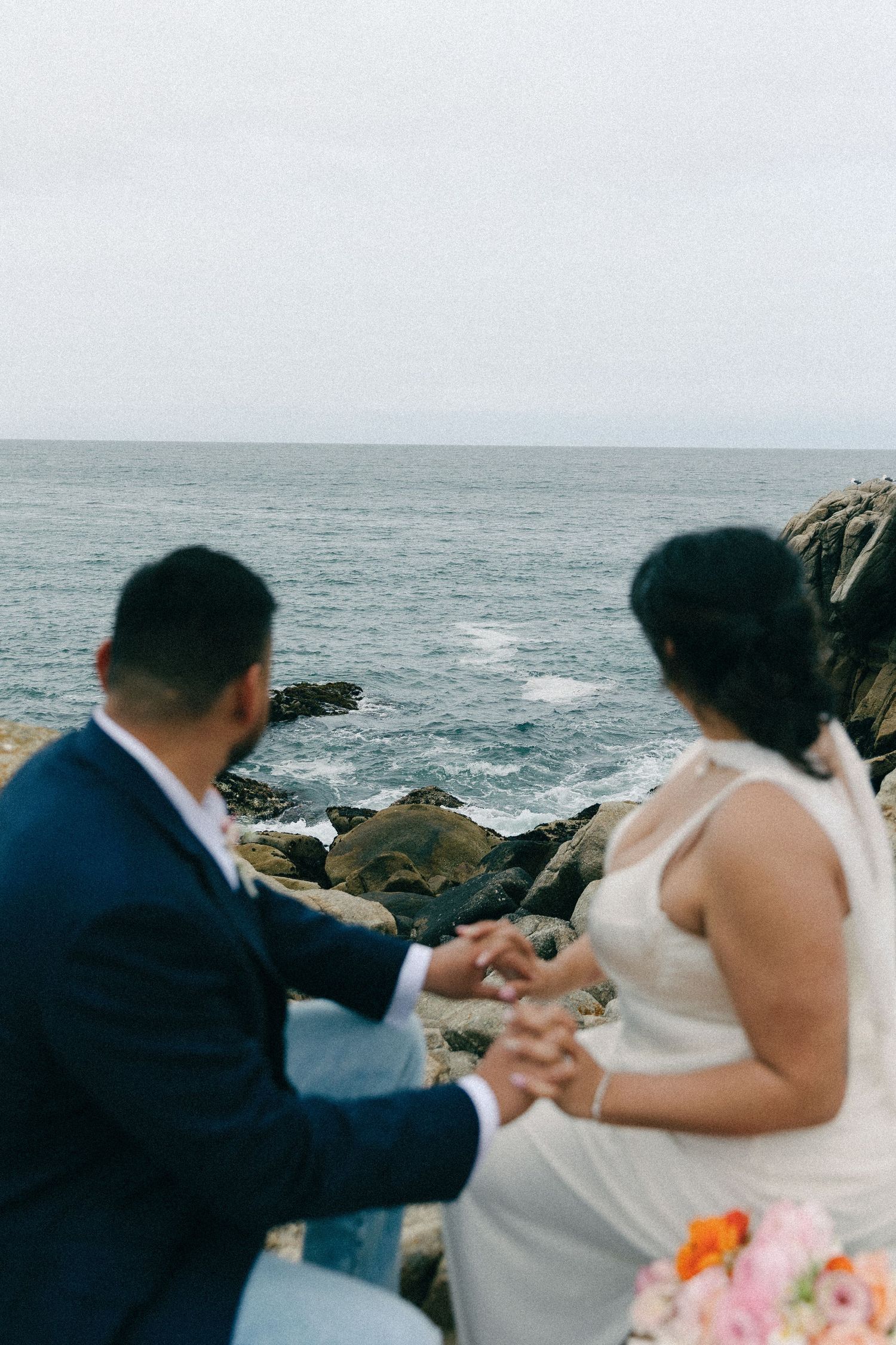 A couple shares an intimate moment on rocky coastal cliffs overlooking the ocean on their wedding day.