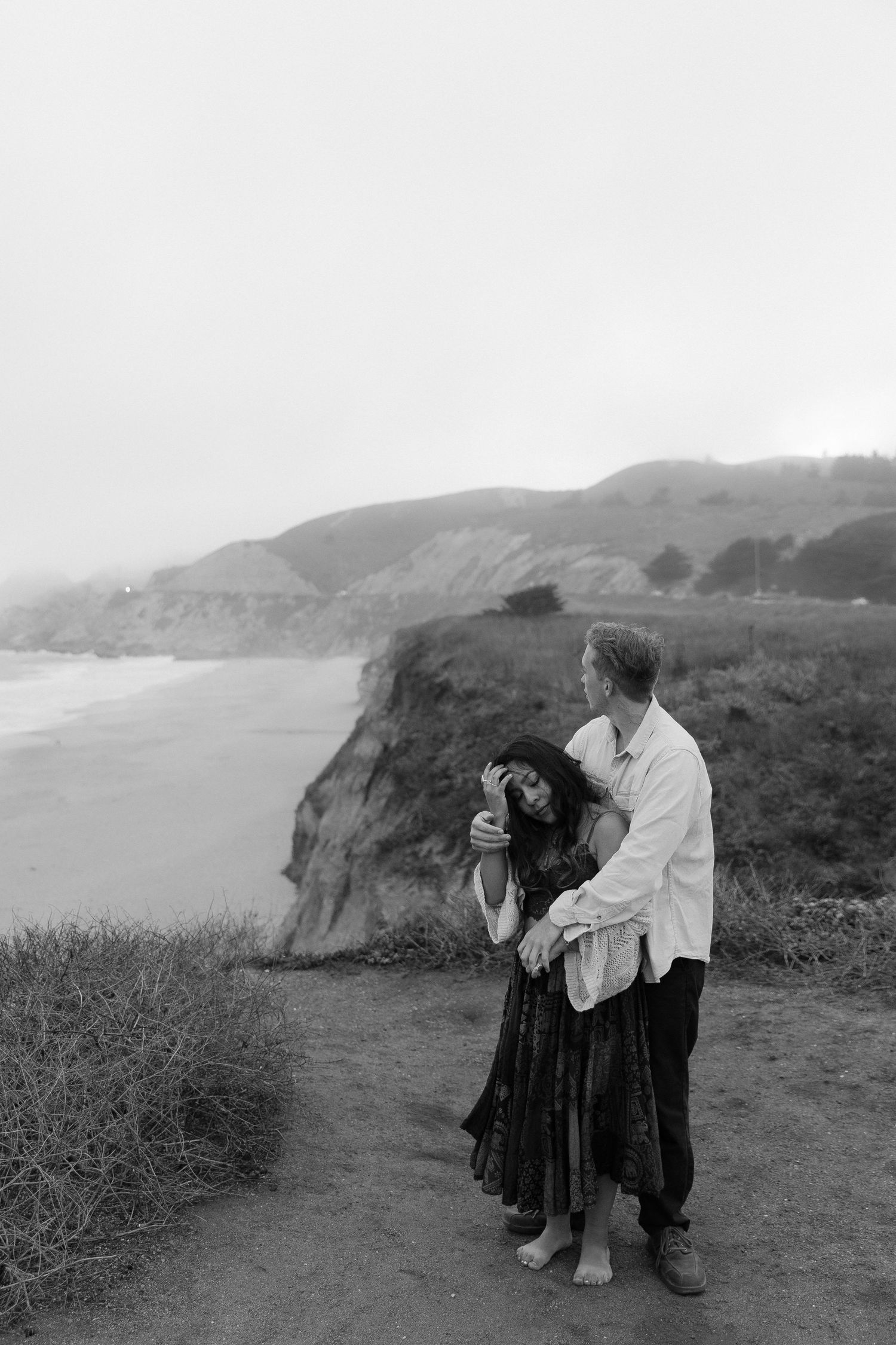 Black and white engagement photo sequence shows couple posing romantically on coastal cliffs overlooking the ocean.