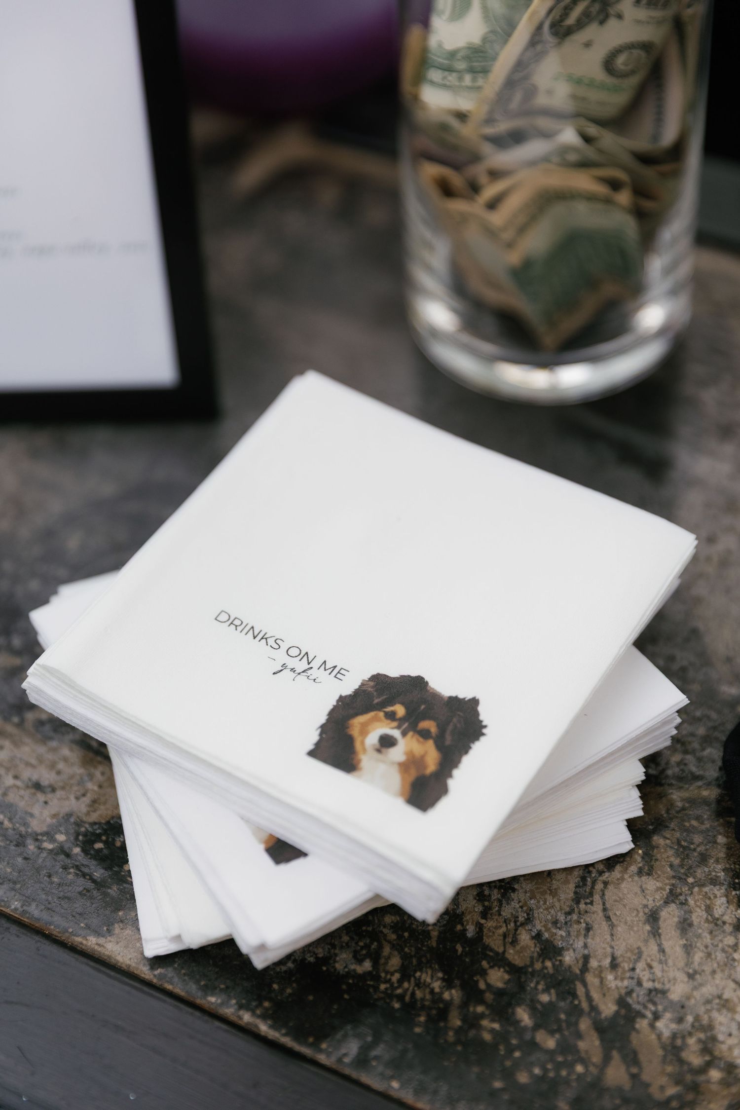 Stack of white napkins with a cute dog illustration on top next to a glass jar with money.