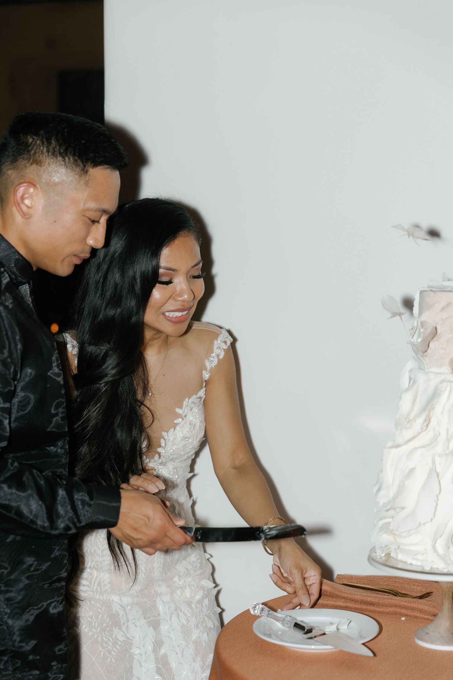 A couple in wedding attire smiles while cutting their elegant white wedding cake together at their reception.