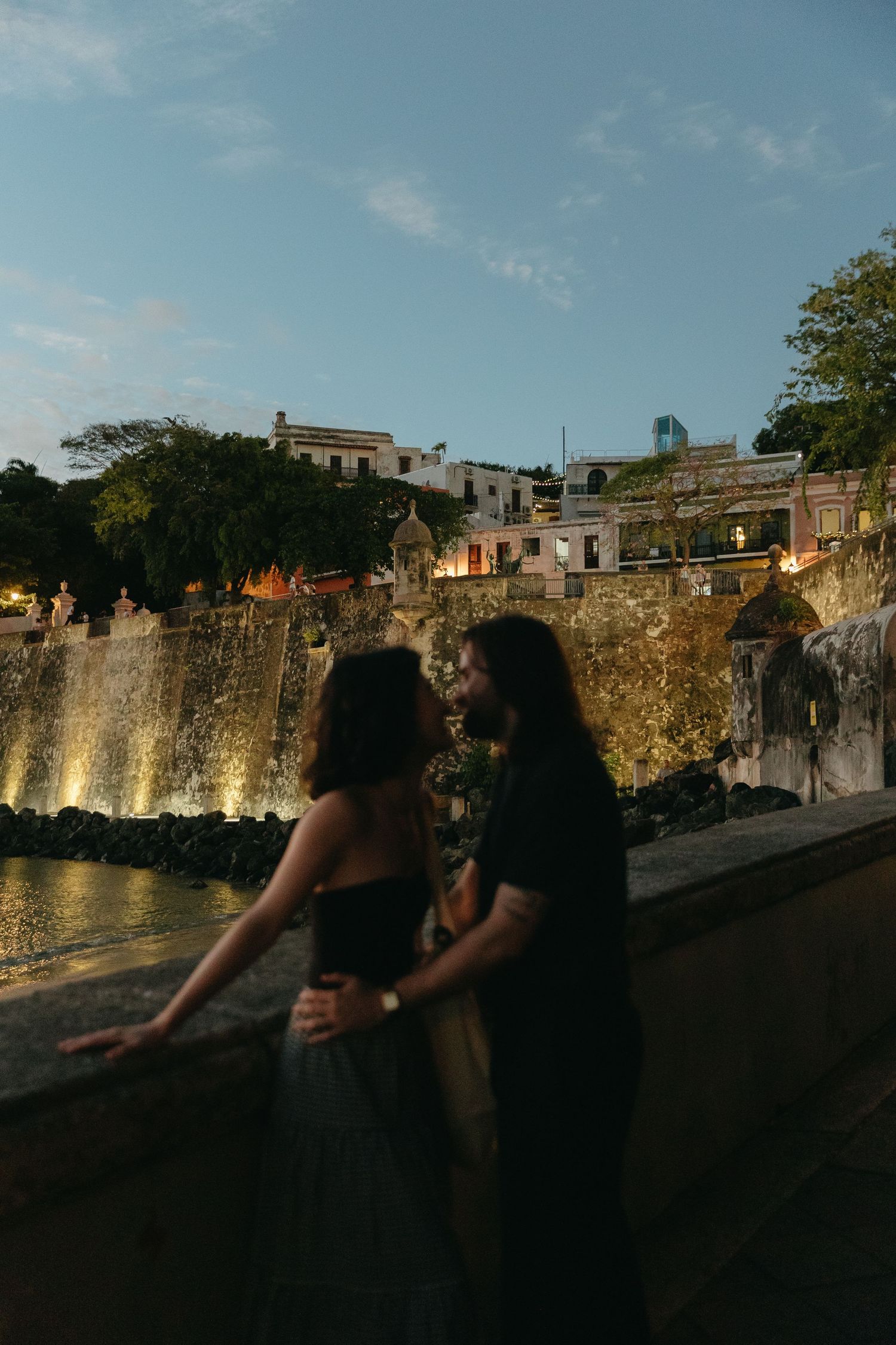 Two silhouettes stand against a stone wall at sunset with buildings visible in the background.