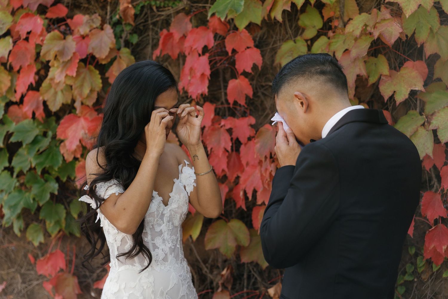 Newlywed couple embraces in front of vibrant red autumn ivy on stone wall during wedding photoshoot.
