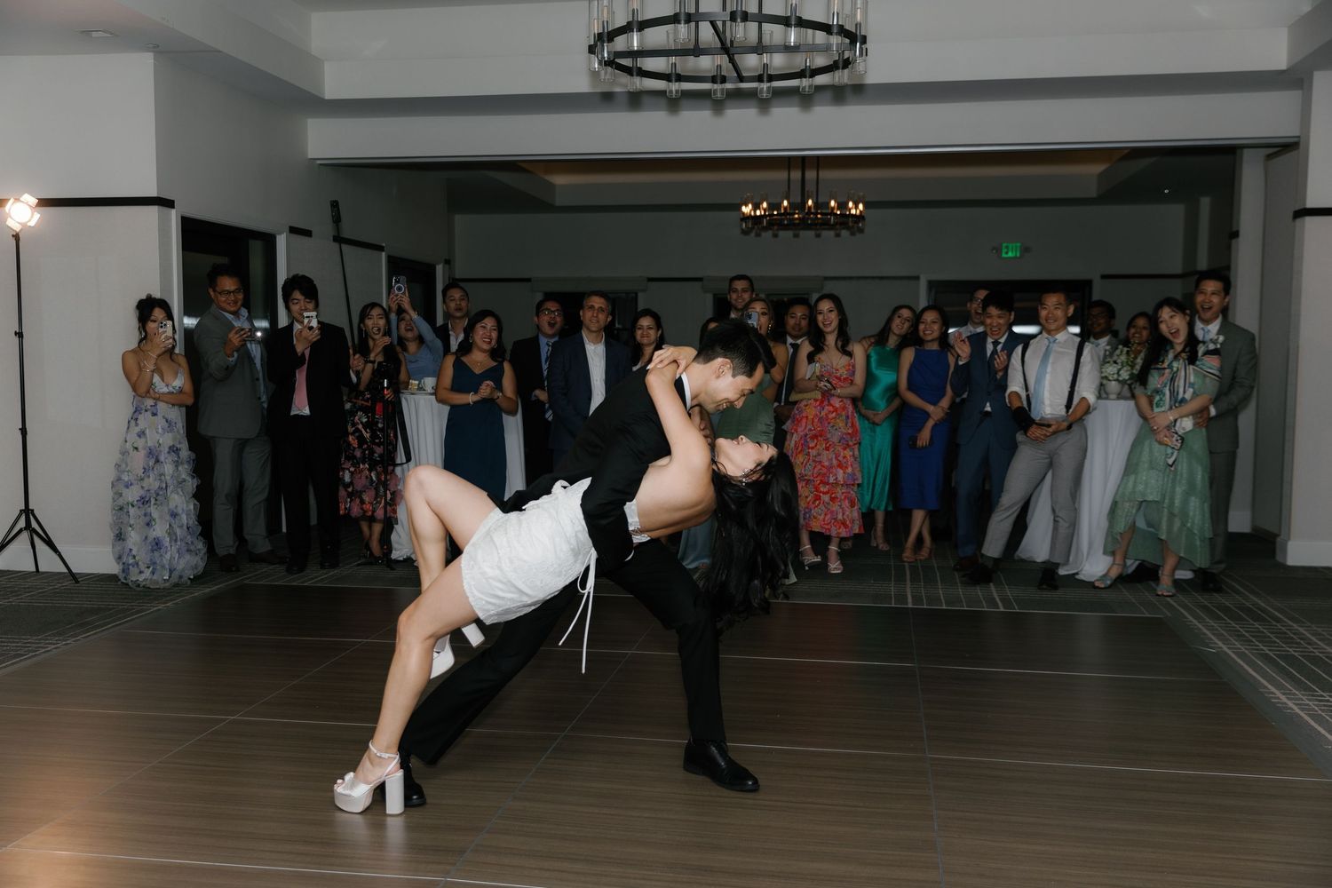 People dancing in a ballroom studio with a chandelier overhead and mirrors on the walls during a dance event.