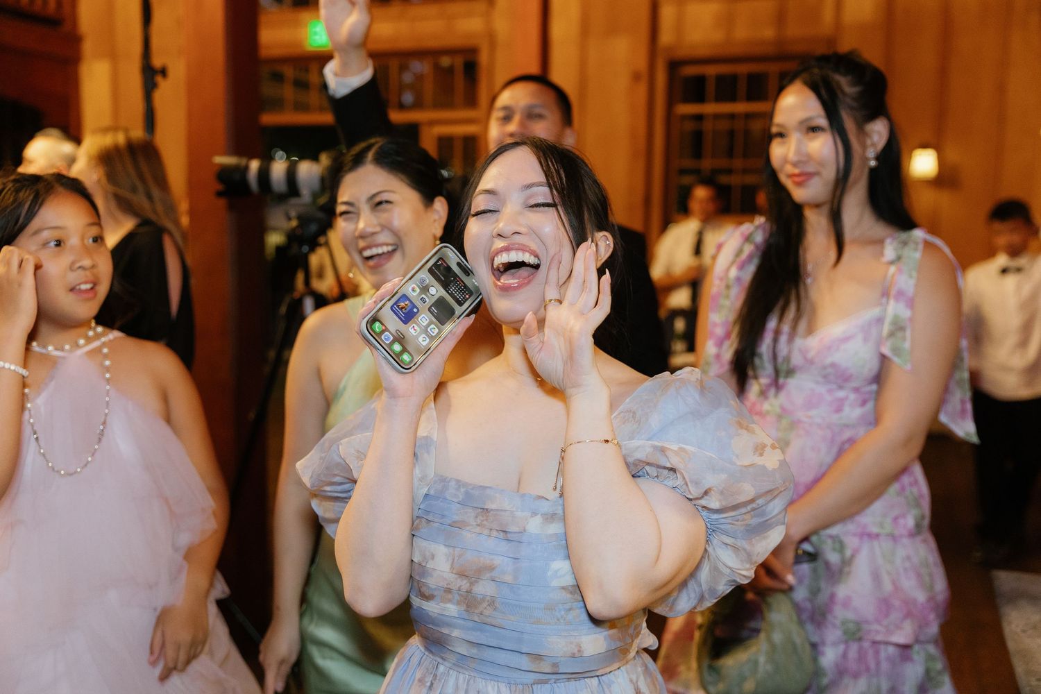 A group of wedding guests in pastel dresses dance and laugh together on the dance floor at a reception.