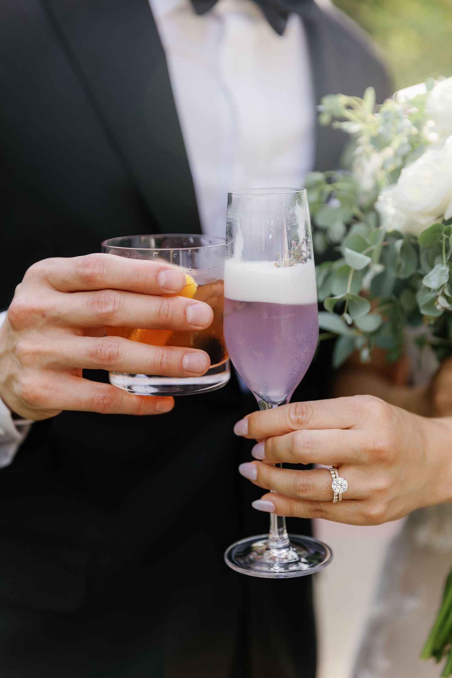 Hands holding purple cocktails with engagement ring visible during a wedding celebration.