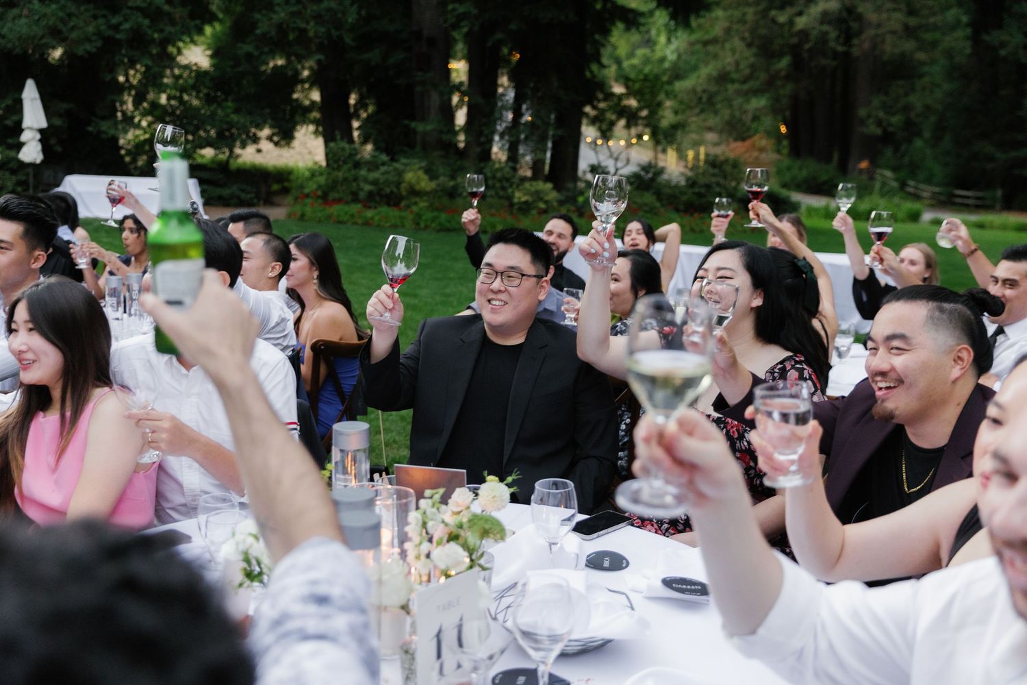 A large group of wedding guests raise their glasses in a celebratory toast during an outdoor reception.