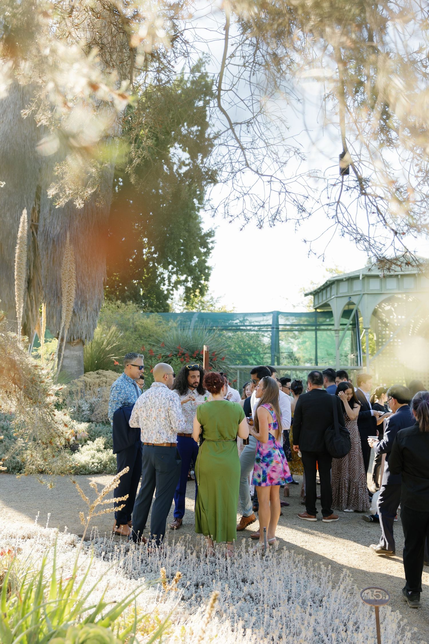 A group of guests gathered outdoors at a garden event with lush greenery and landscaping in the background.