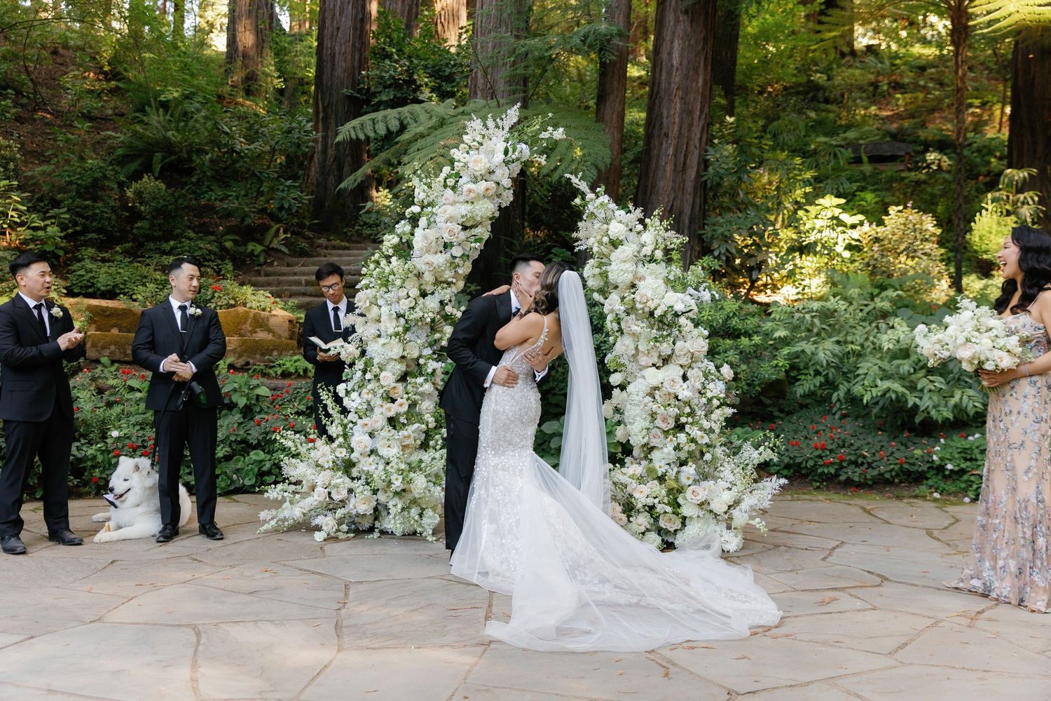 A wedding ceremony takes place under an elegant white floral arch in a serene forest setting.