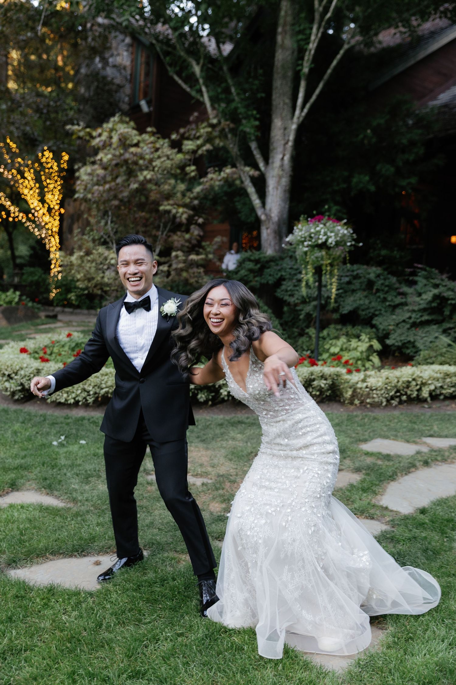 A joyful wedding couple poses playfully on grass with trees and string lights in the background.