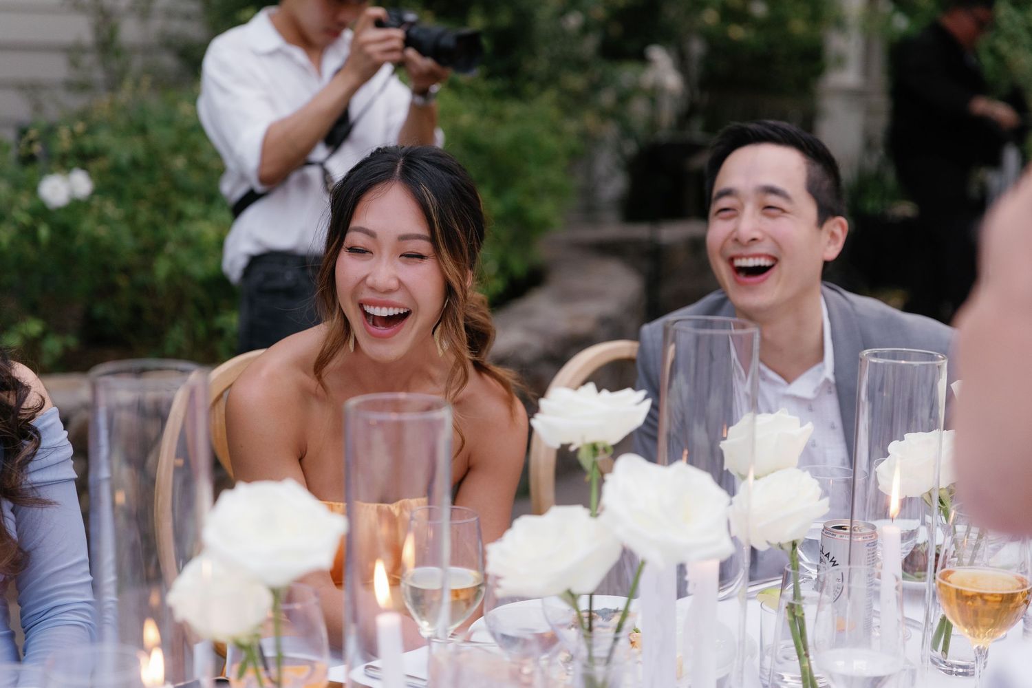 Wedding guests seated at an outdoor reception table with white floral centerpieces and glassware.