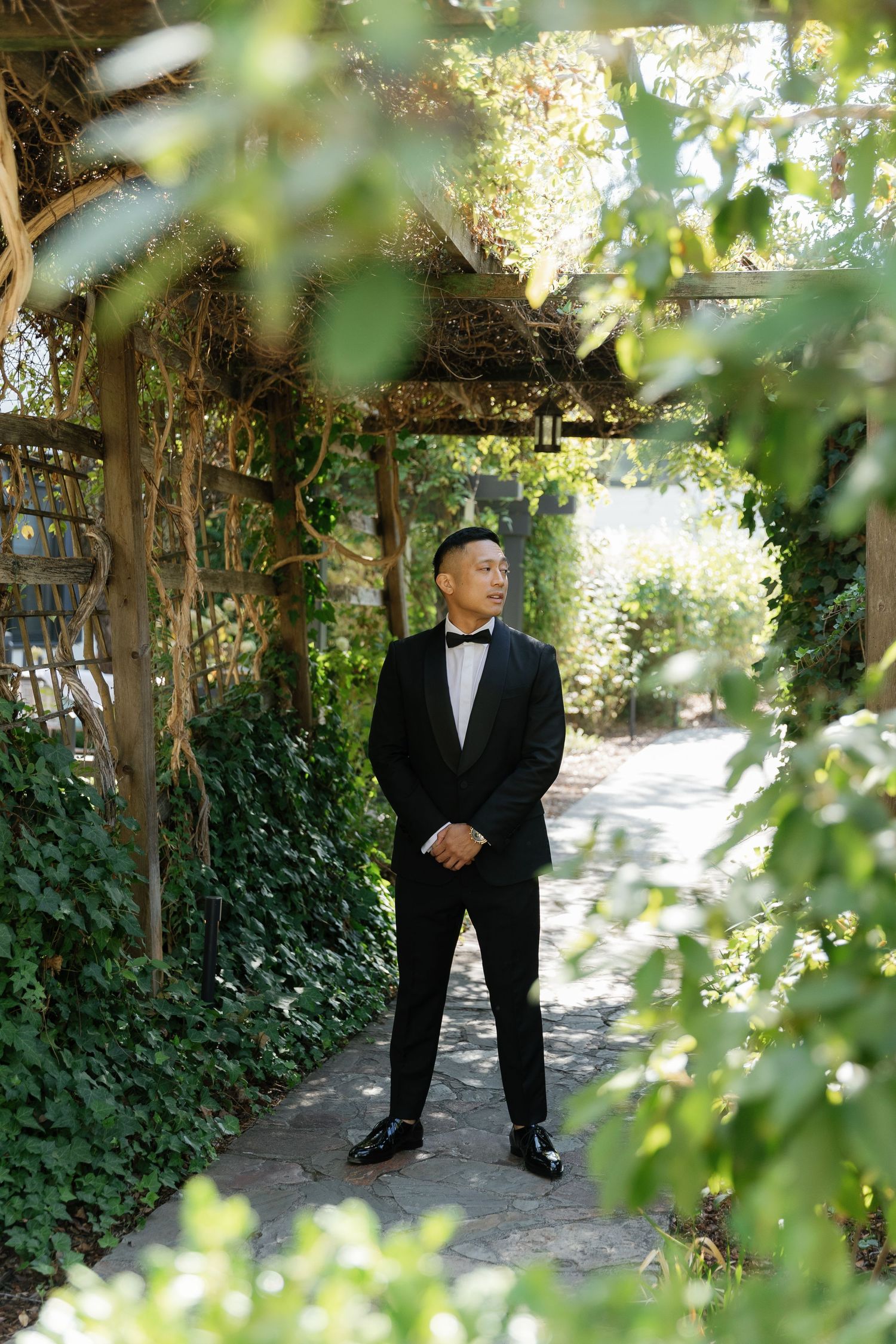 A person in a formal black tuxedo stands on a garden path surrounded by lush green foliage and trees.