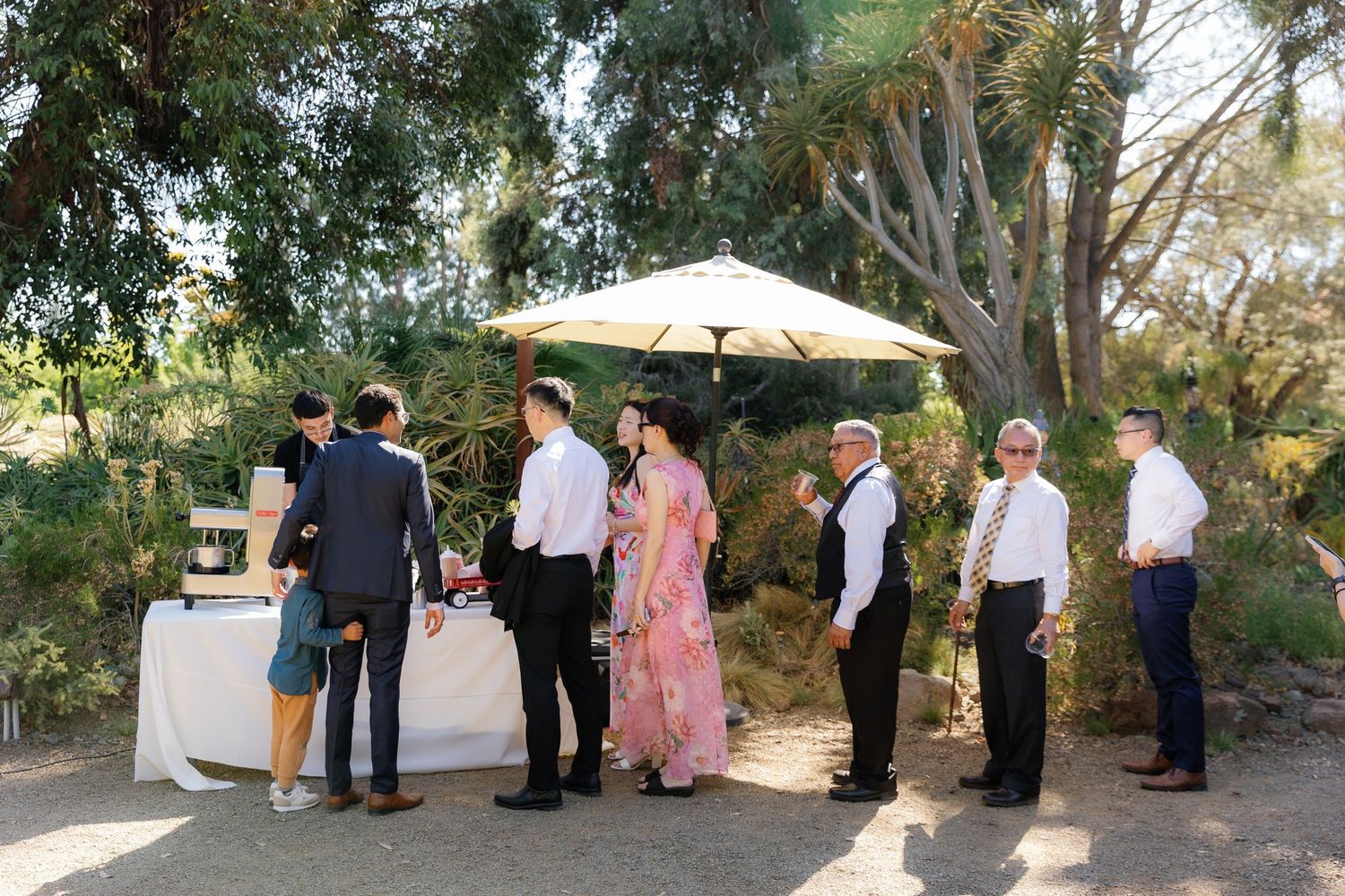Wedding guests gather outdoors under trees and a white umbrella during a garden ceremony.