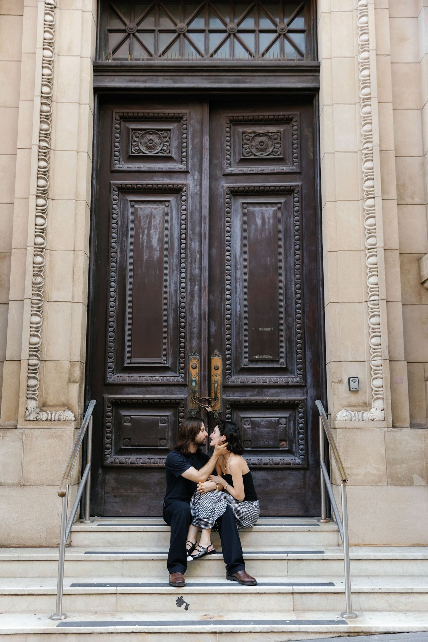 A couple sits intimately on steps in front of large ornate wooden doors on a historic building.