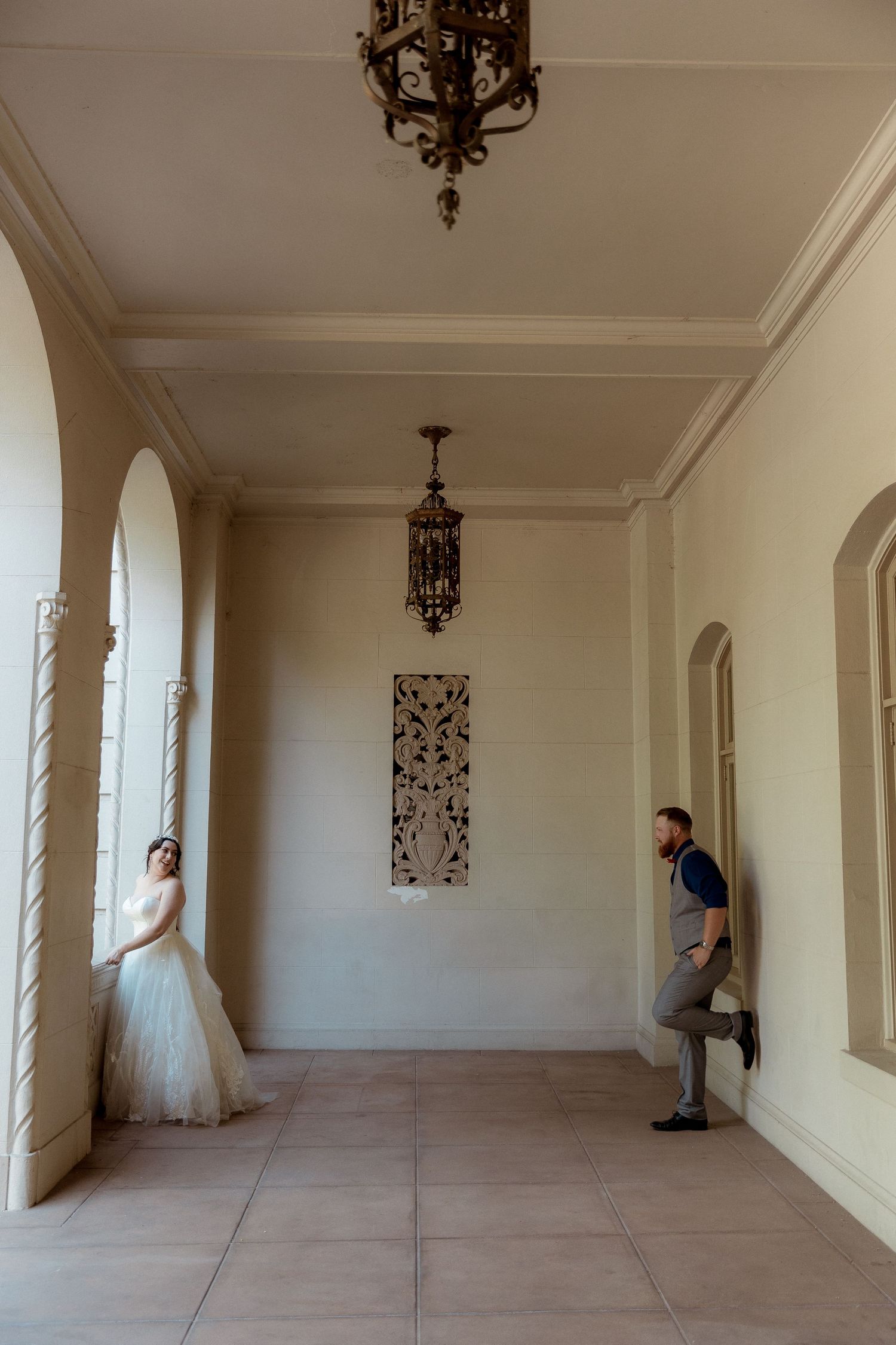 Two people in wedding attire pose playfully in an elegant hallway with ornate chandeliers.